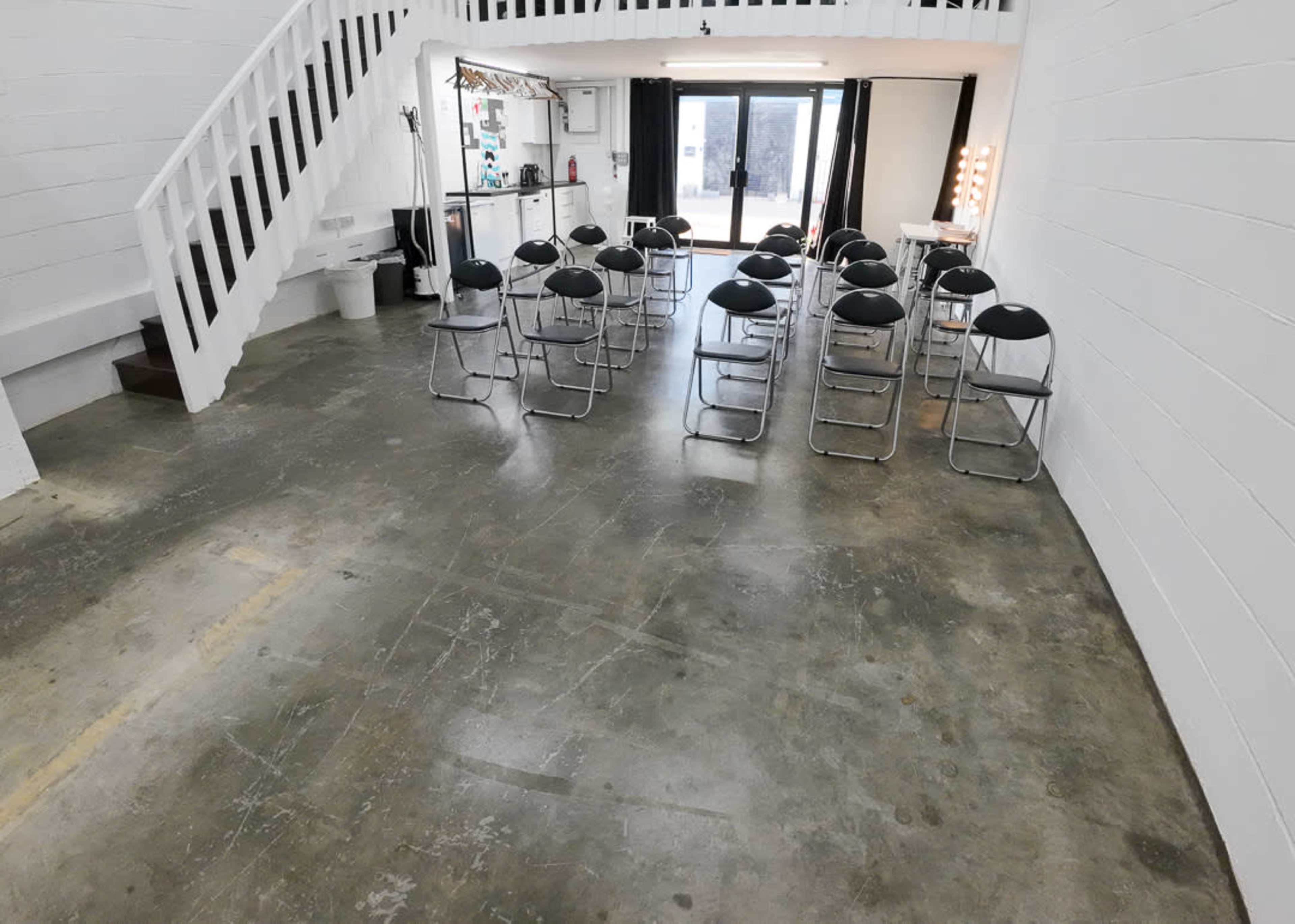 A large, empty room with a polished concrete floor and a row of black folding chairs arranged in the center, adjacent to a staircase and large windows.