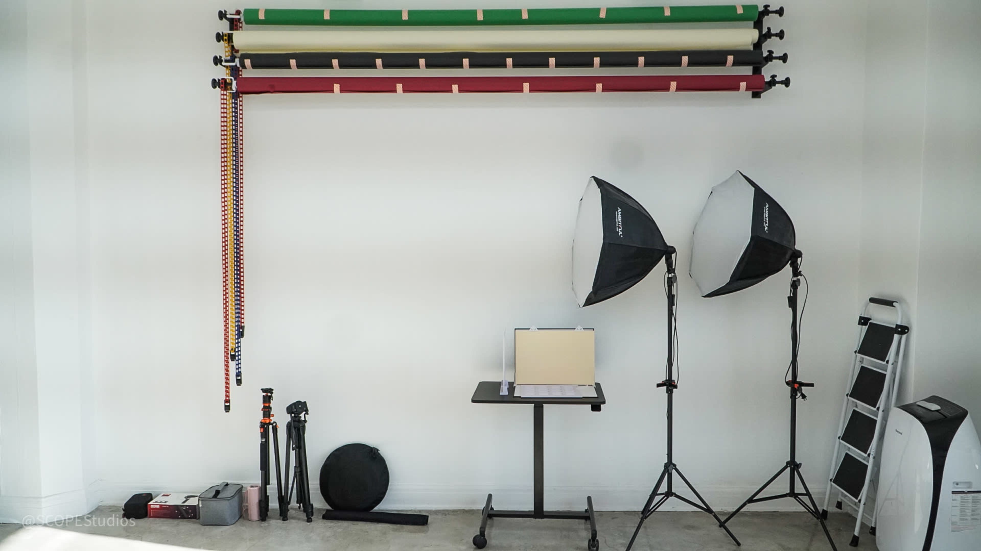 The image shows a photography studio setup with backdrop rolls hung on the wall, two softbox lights, a table with a light reflector, and various camera equipment positioned nearby.