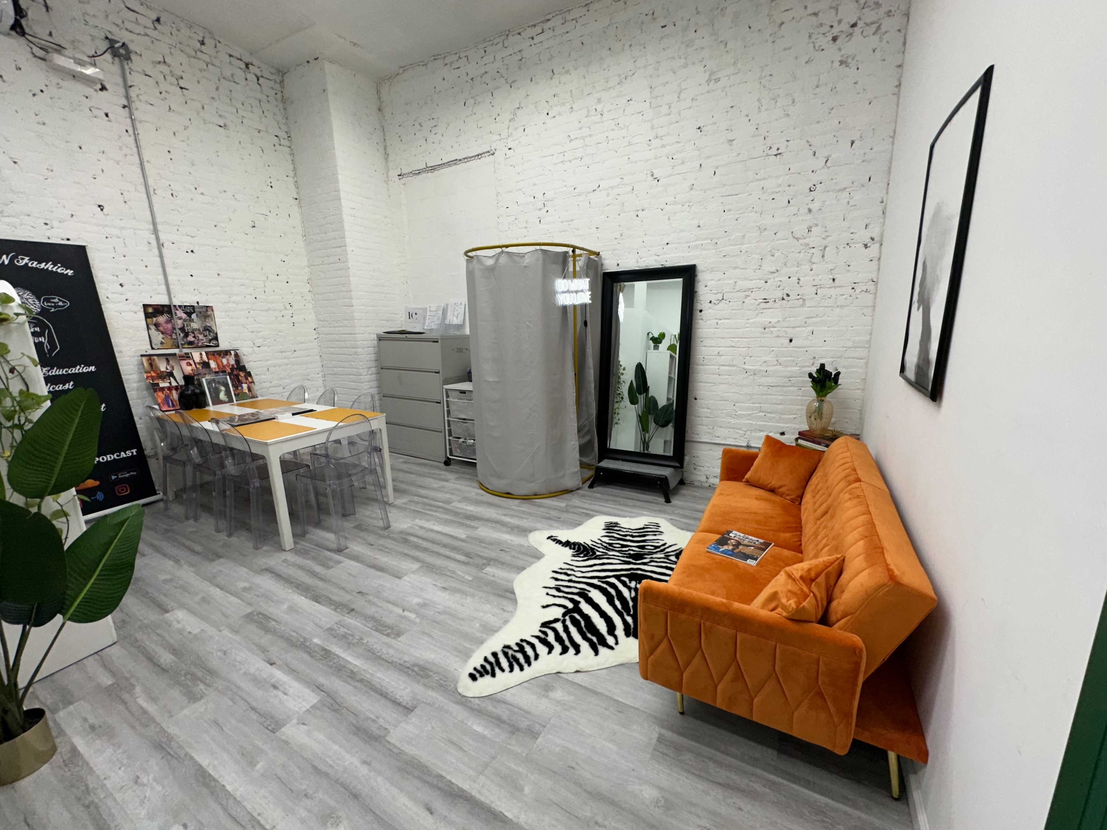 A modern office space features an orange sofa, a glass dining table, a clothing rack, and potted plants against a backdrop of white brick walls.