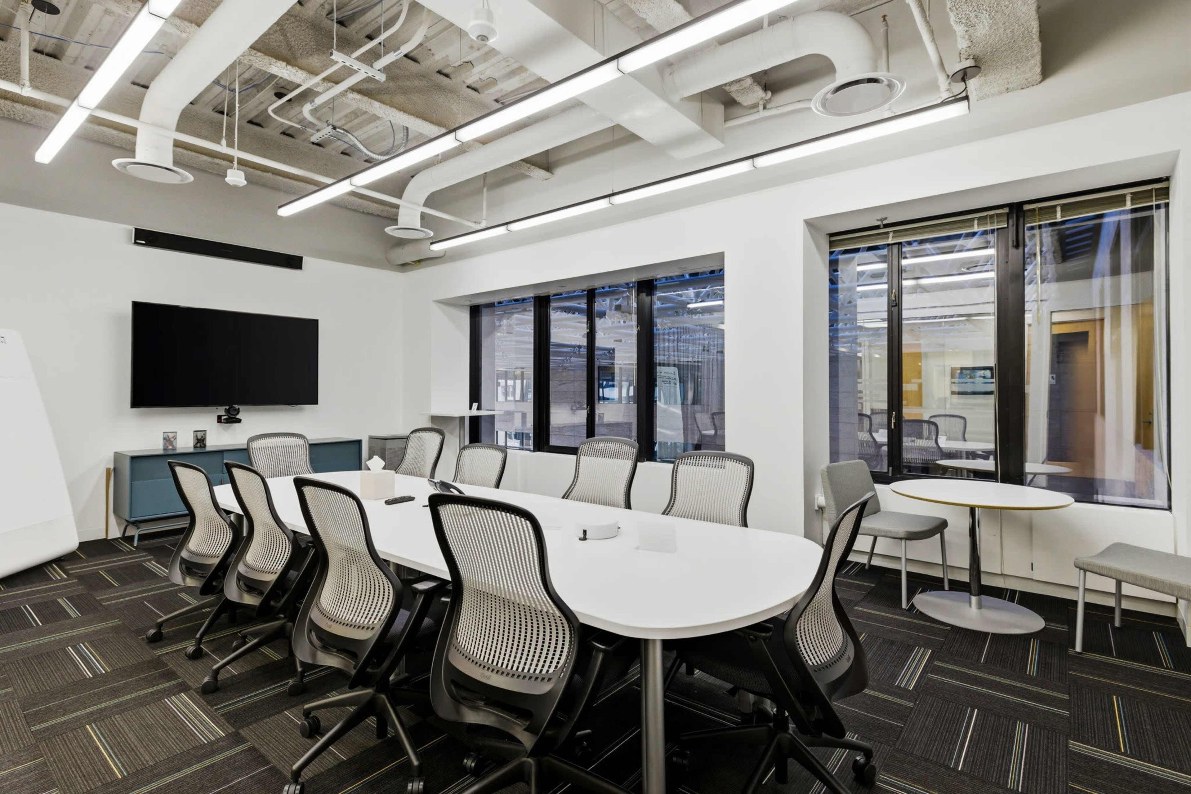 The image shows a modern conference room featuring a long, white oval table surrounded by ergonomic chairs, with large windows allowing natural light and a flat-screen TV mounted on the wall.