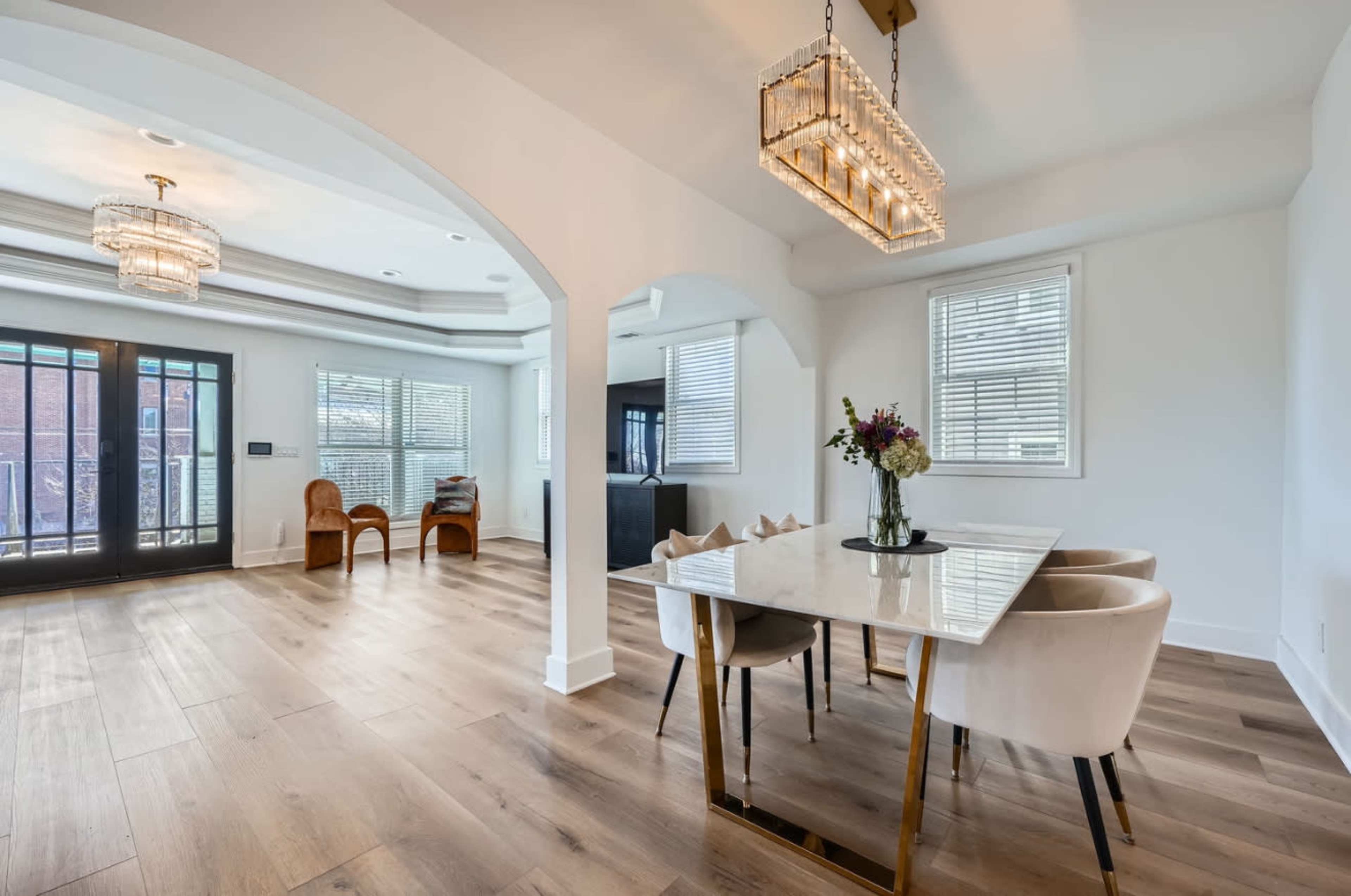 The image shows a bright dining area featuring a marble-topped table with upholstered chairs, adjacent to a living space with large windows and modern lighting fixtures.