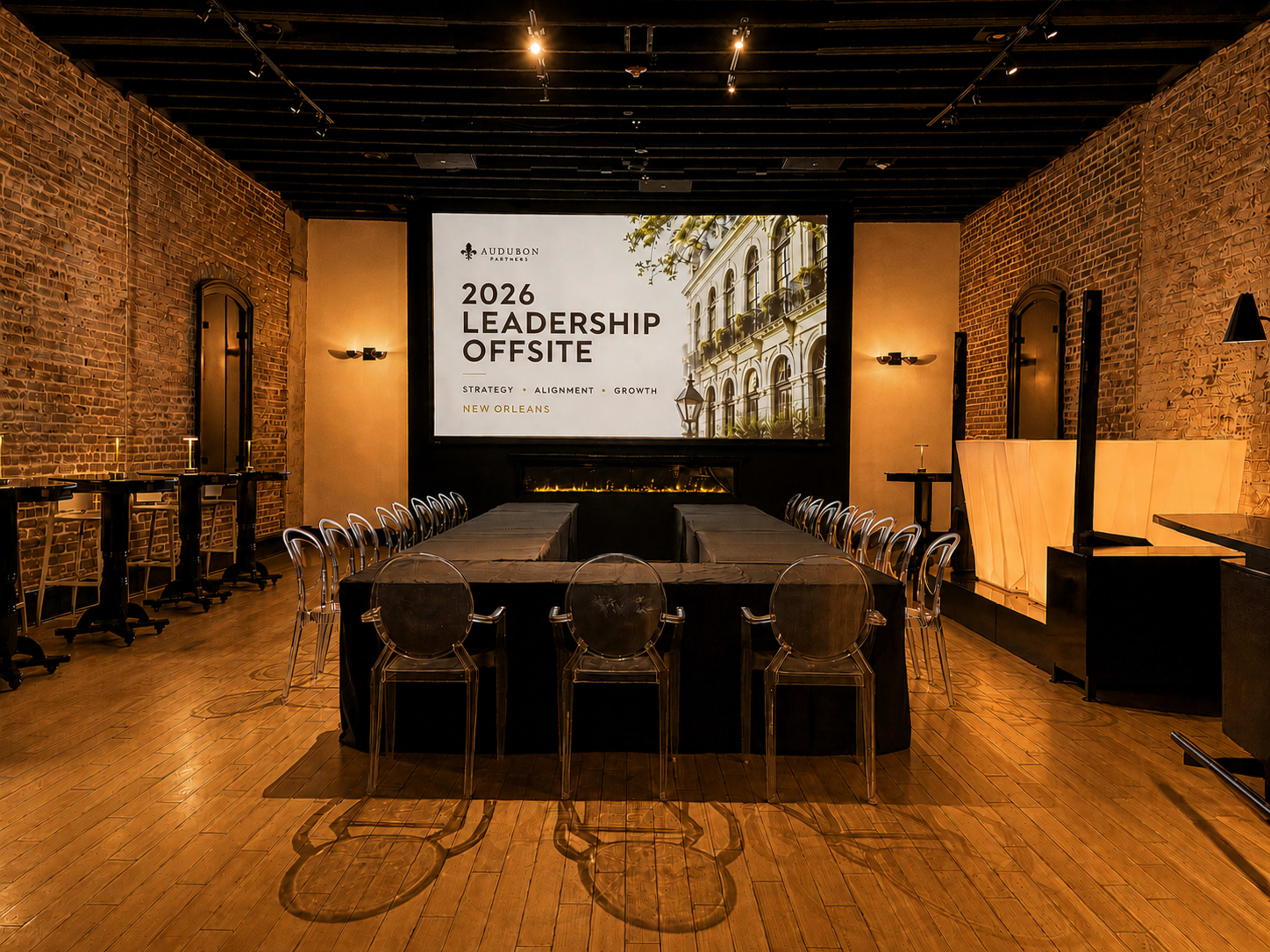 The image shows a conference room with a large screen displaying "2026 Leadership Offsite" and chairs arranged around a central table in a rustic setting with exposed brick walls.