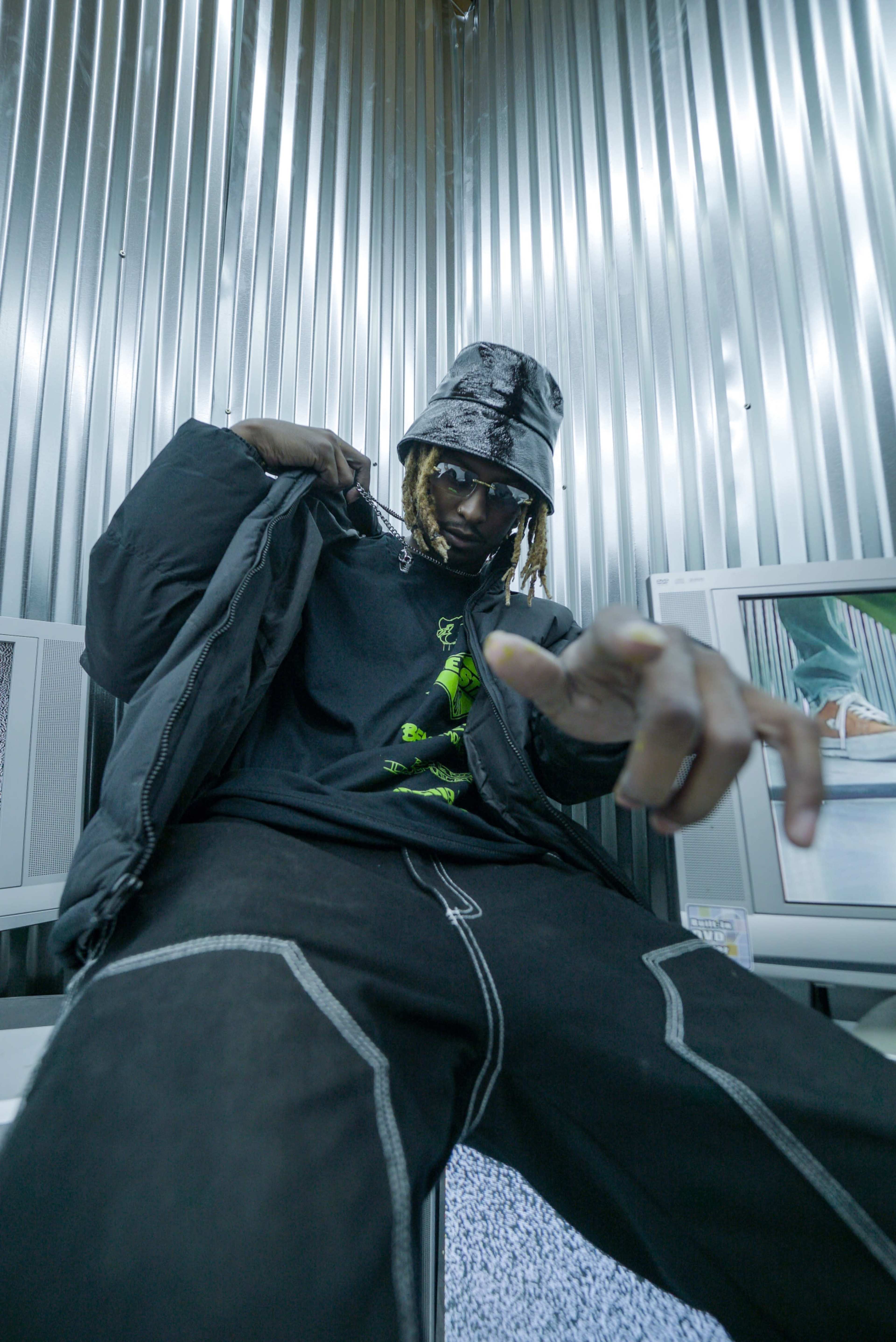 A person wearing a black bucket hat and oversized jacket poses in a metallic room with old televisions around them.