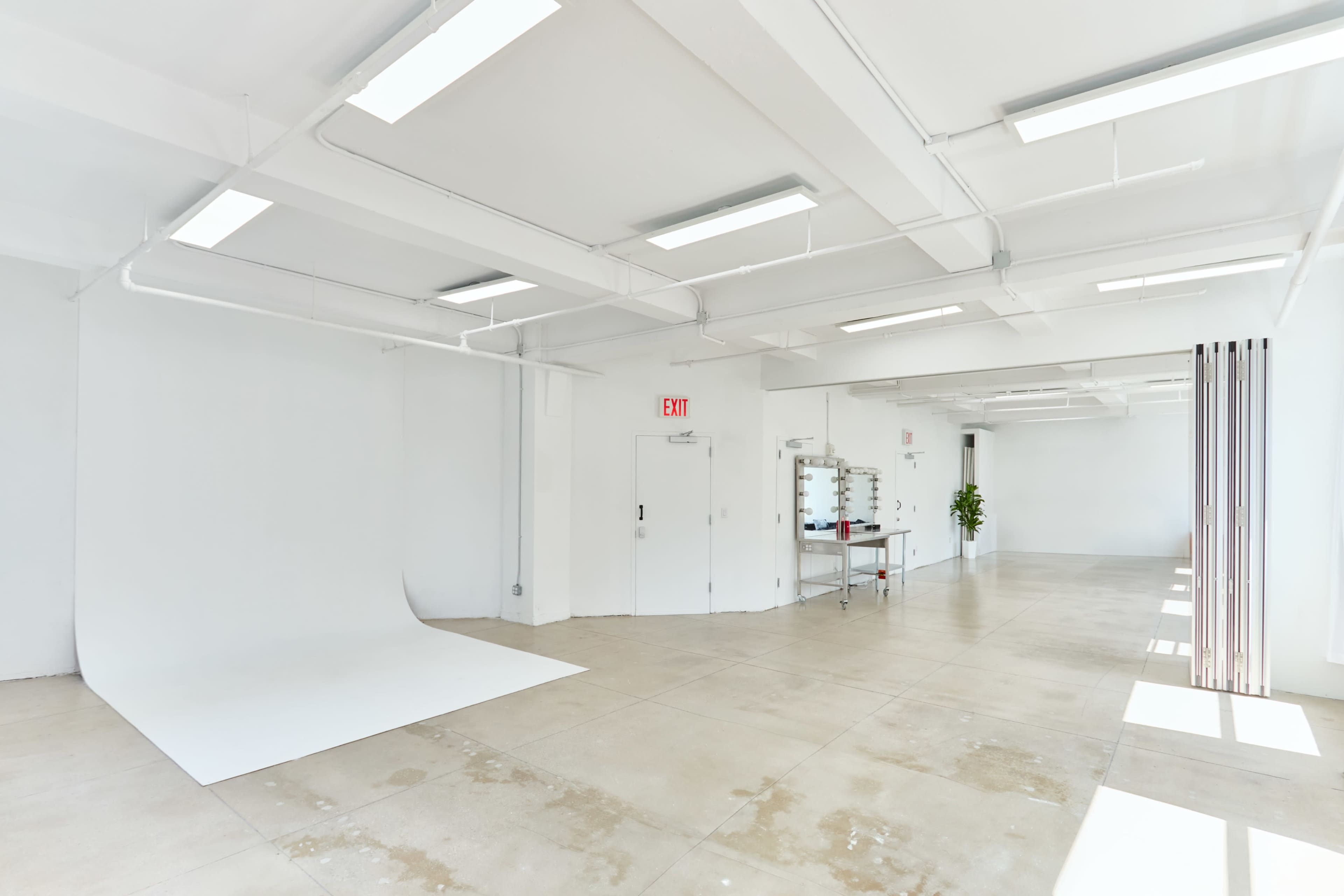 The image shows a spacious, empty studio with white walls, concrete flooring, a backdrop area, and a feature with a mirror and a plant in one corner.