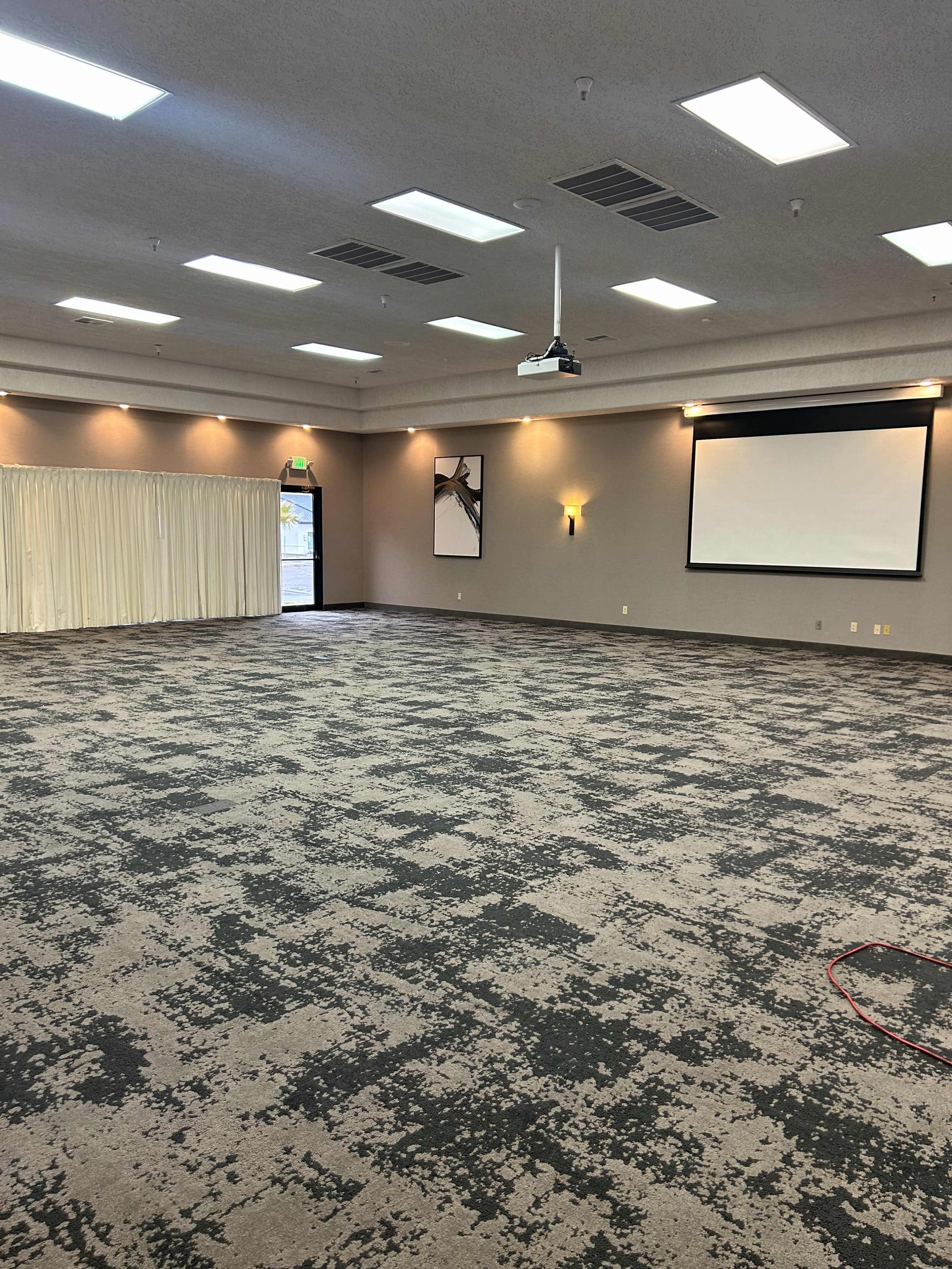 The image shows a spacious, empty conference room with a projector screen on one wall and light-colored curtains covering a window.