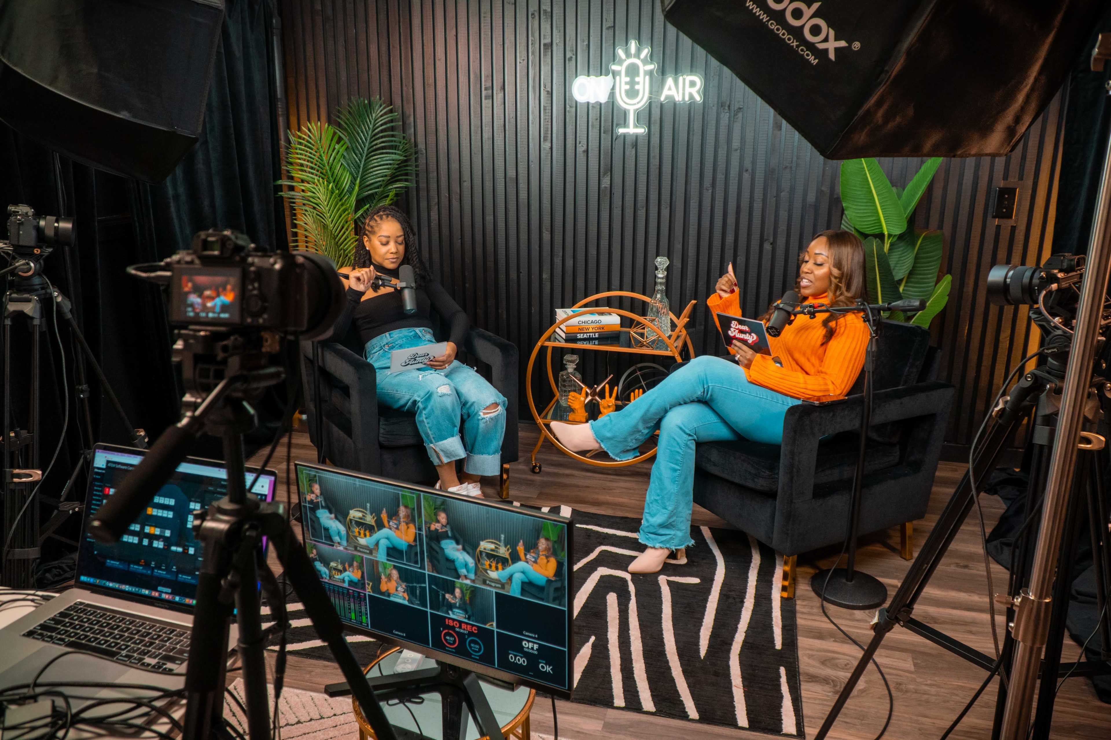 Two women sit in a studio, discussing a topic while surrounded by cameras and production equipment.