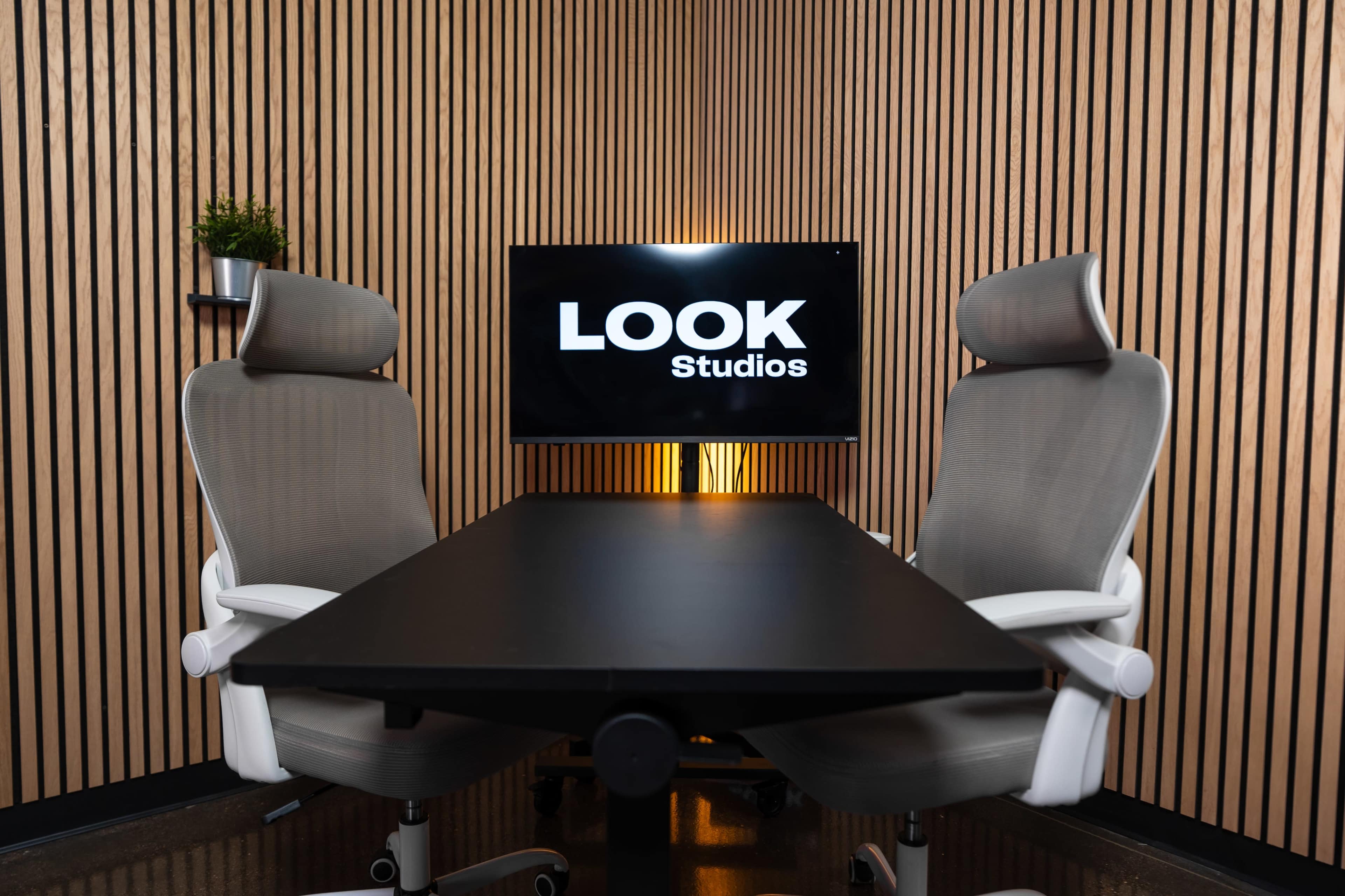 A meeting room features two ergonomic chairs facing a black table, with a television displaying the logo "LOOK Studios" on a wood-paneled wall.