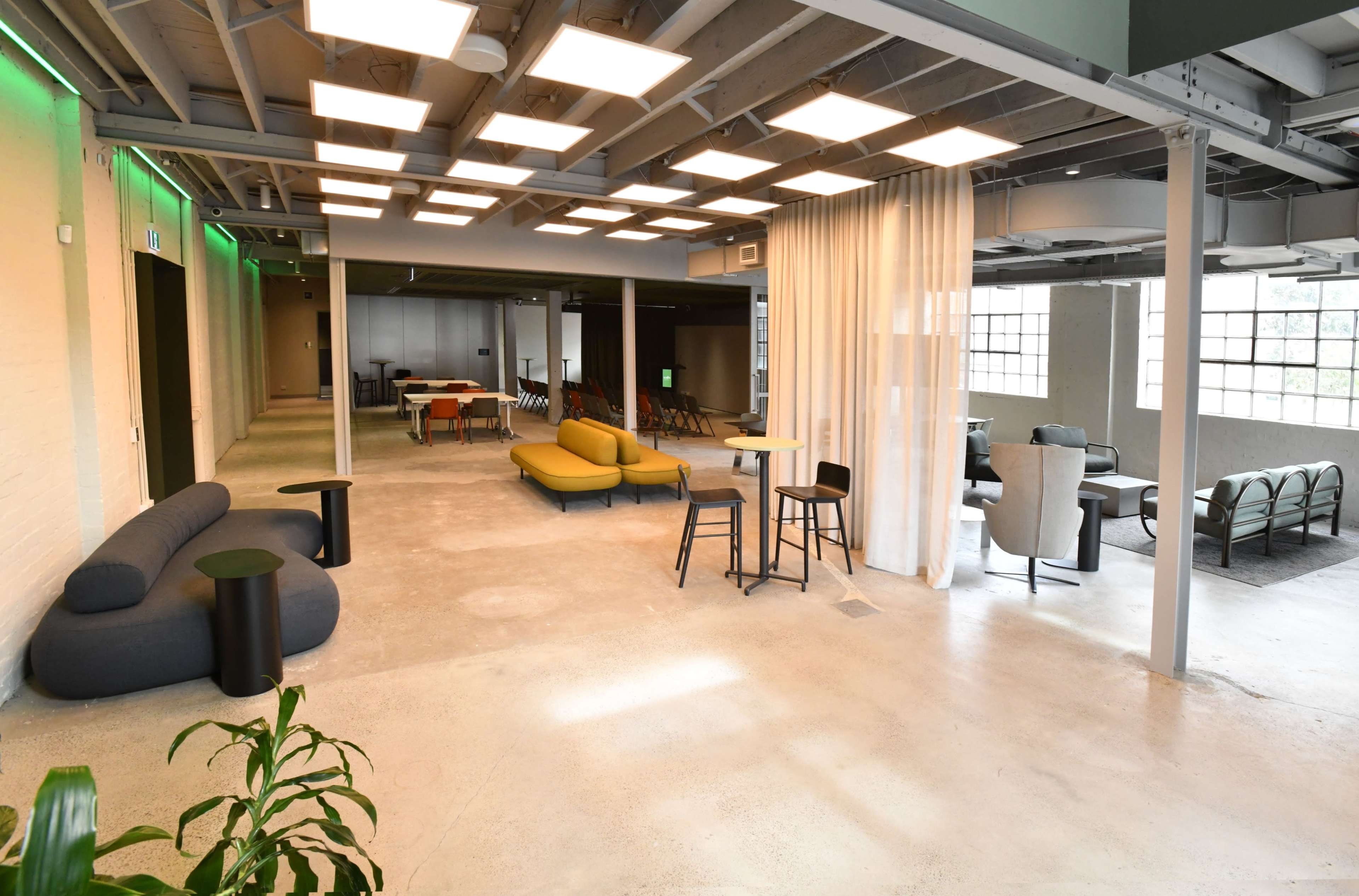 The image shows a spacious, modern interior of an office or co-working space with various seating areas, including sofas and high tables, under bright overhead lighting.