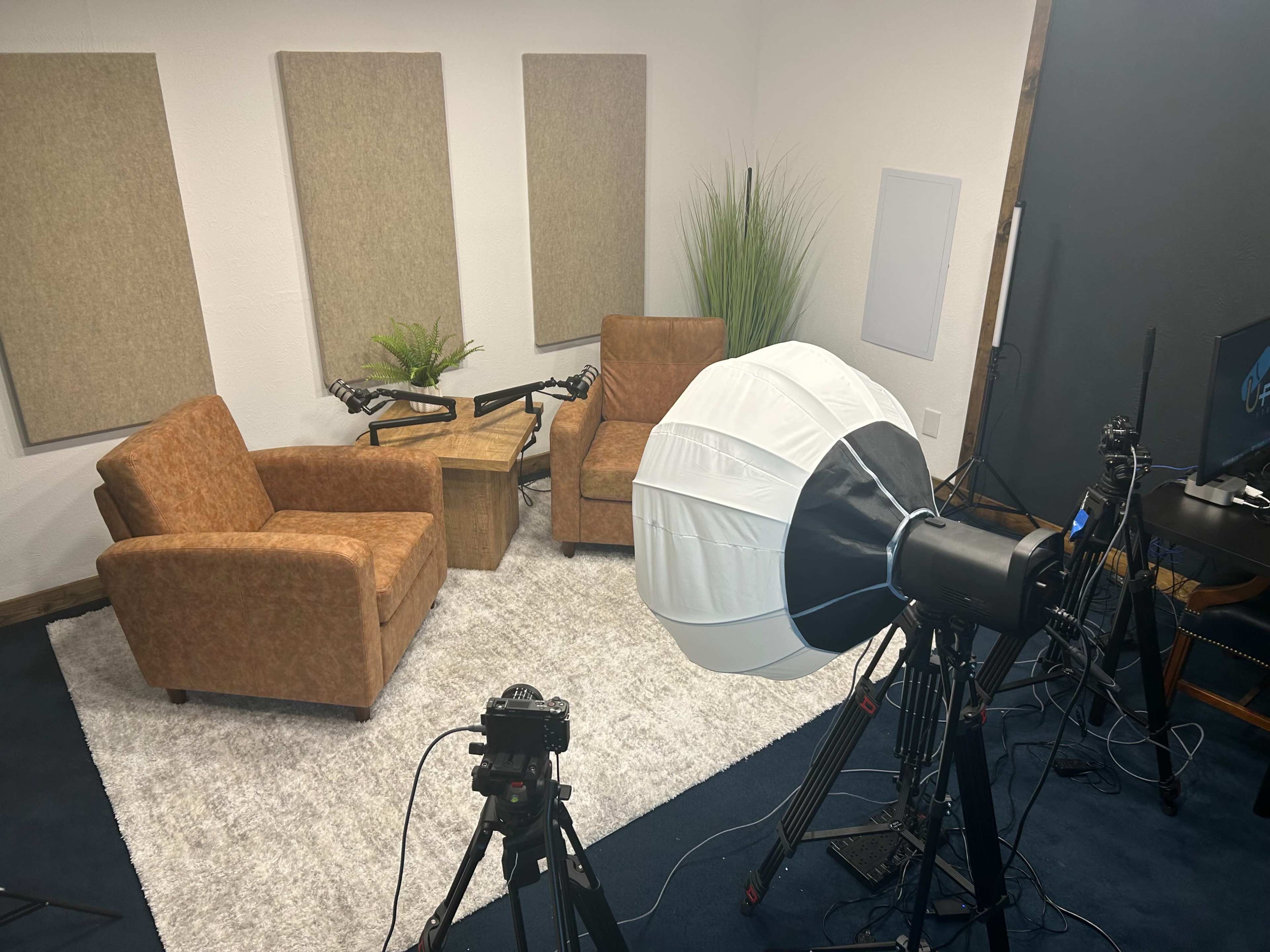 The image shows a small studio setup with two brown armchairs facing a wooden table, surrounded by soft paneling and a large light source.