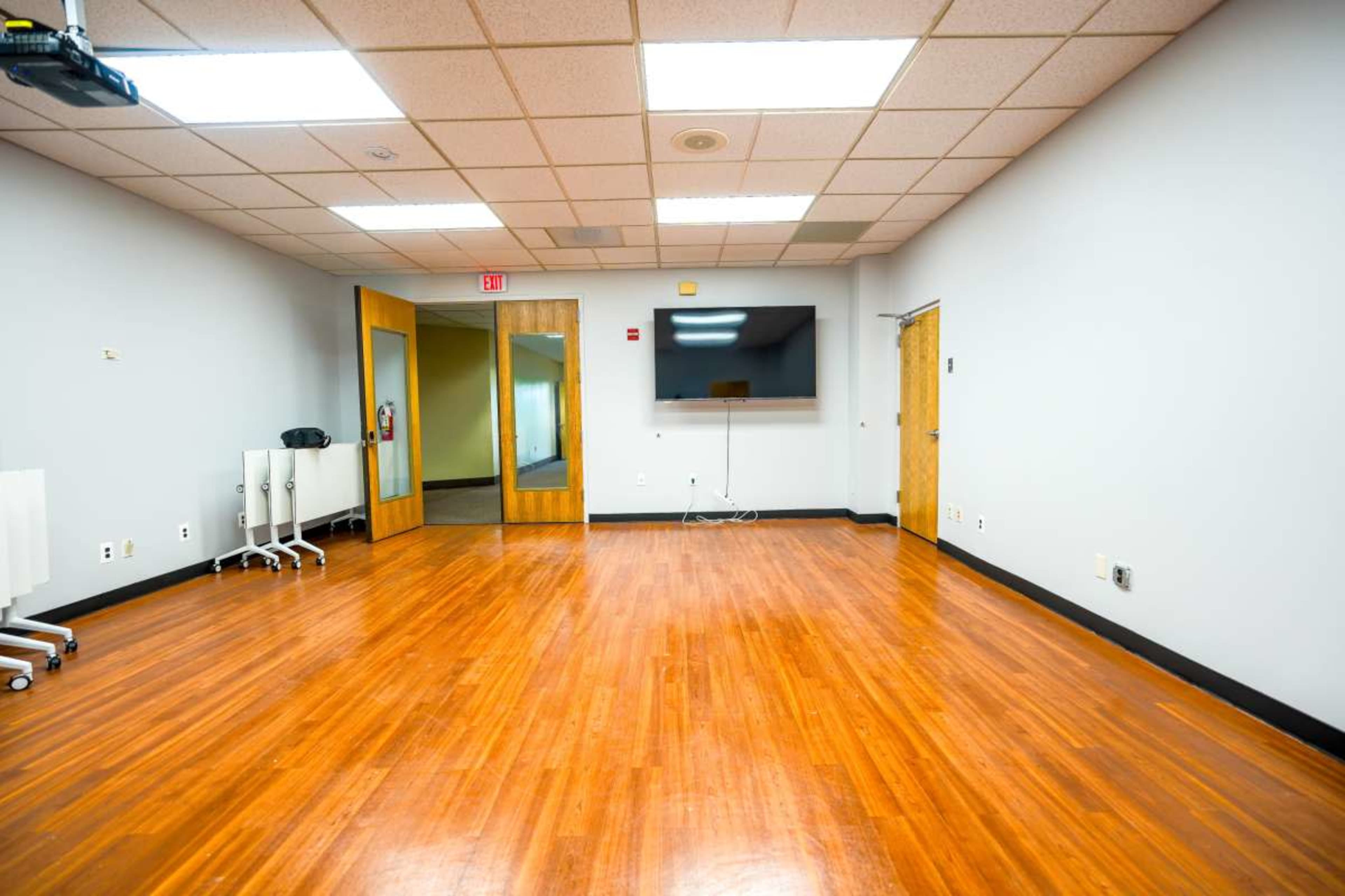 The image shows a spacious, empty meeting room with wooden floors, a wall-mounted television, and two doors leading to other areas.