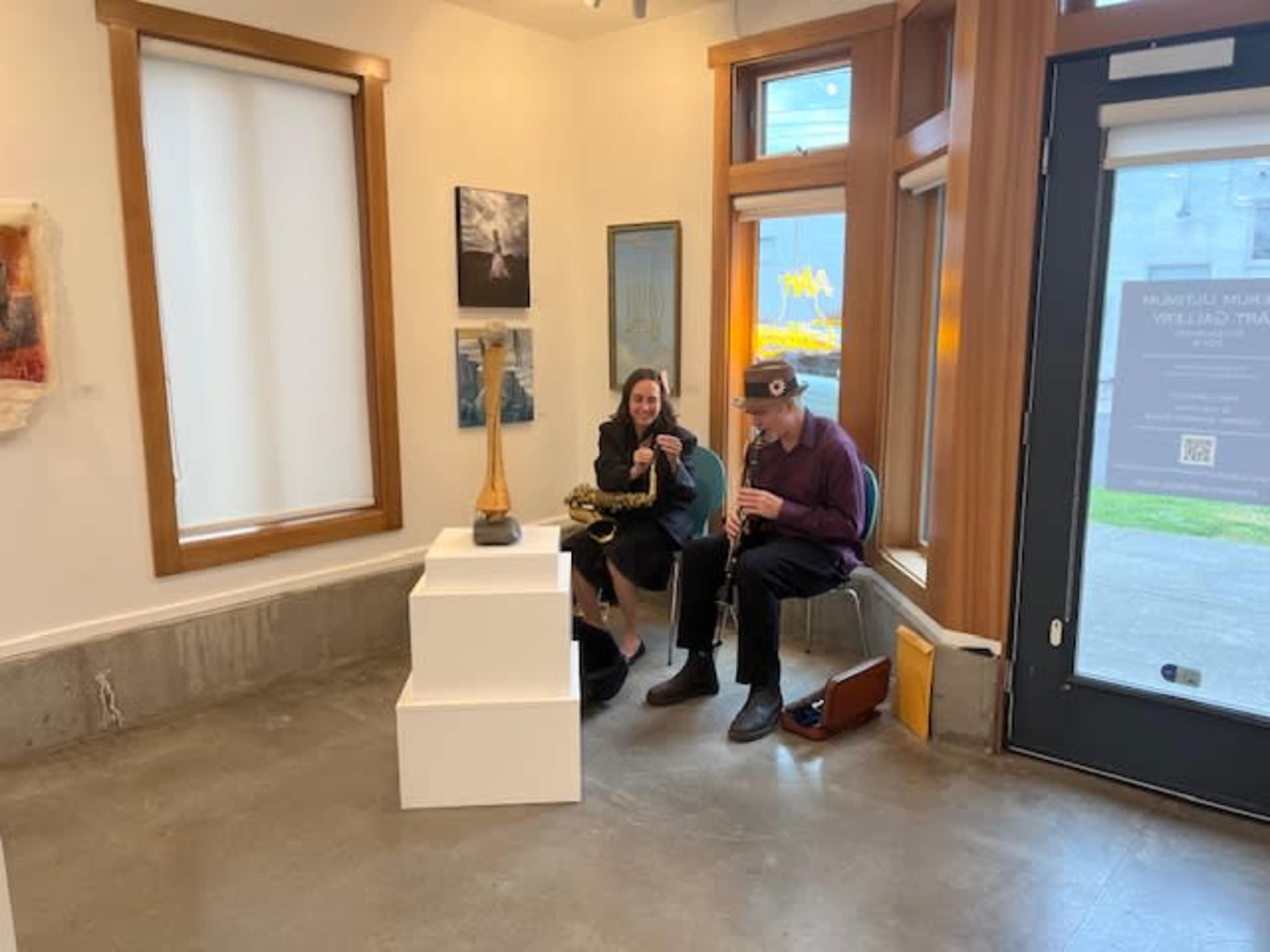 Two musicians sit in a gallery space, one holding a saxophone and the other adjusting a brass instrument, surrounded by artwork on the walls.