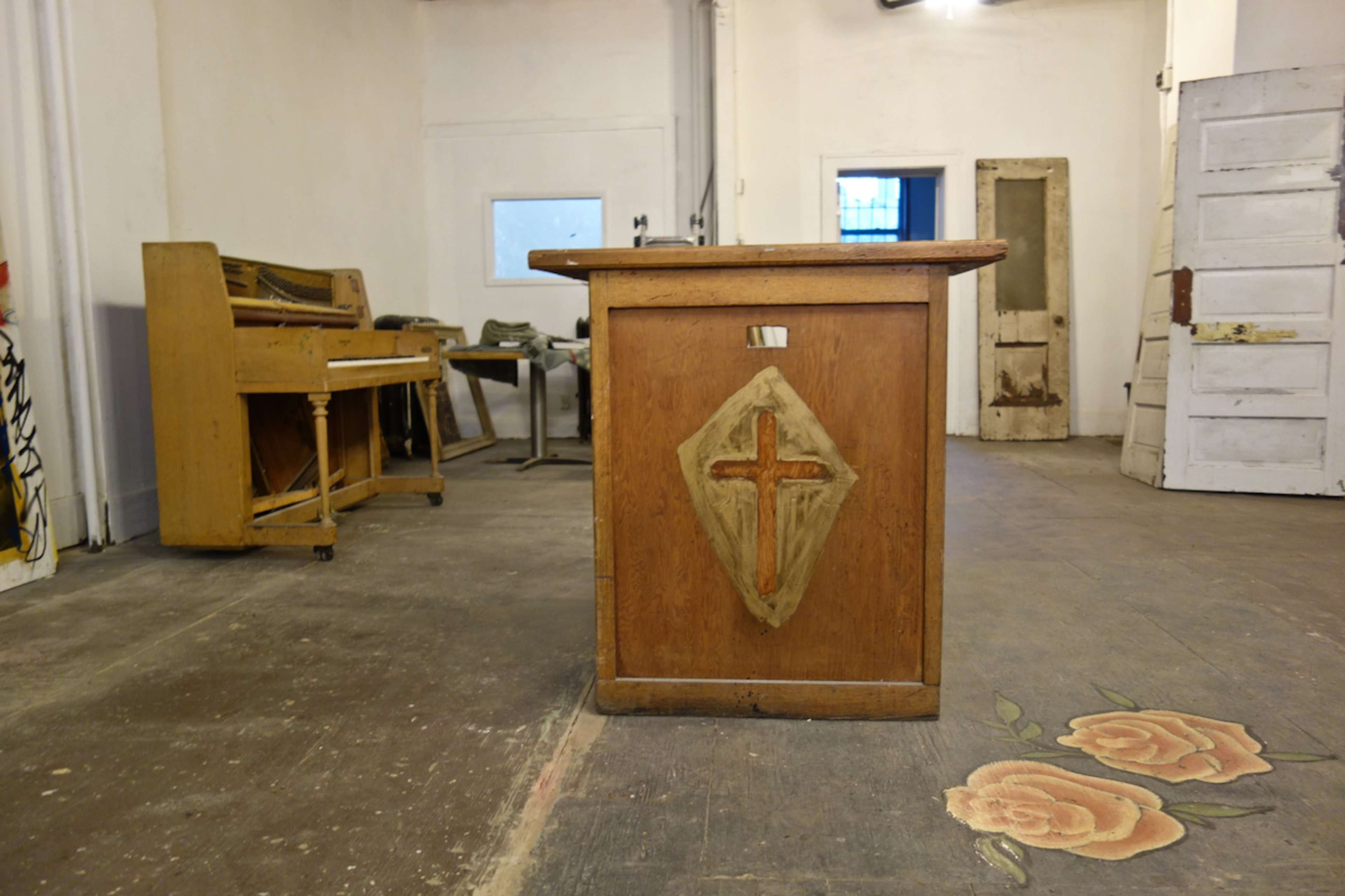 A wooden altar with a cross design in the foreground, set within a sparsely furnished room that includes a piano and a door in the background.