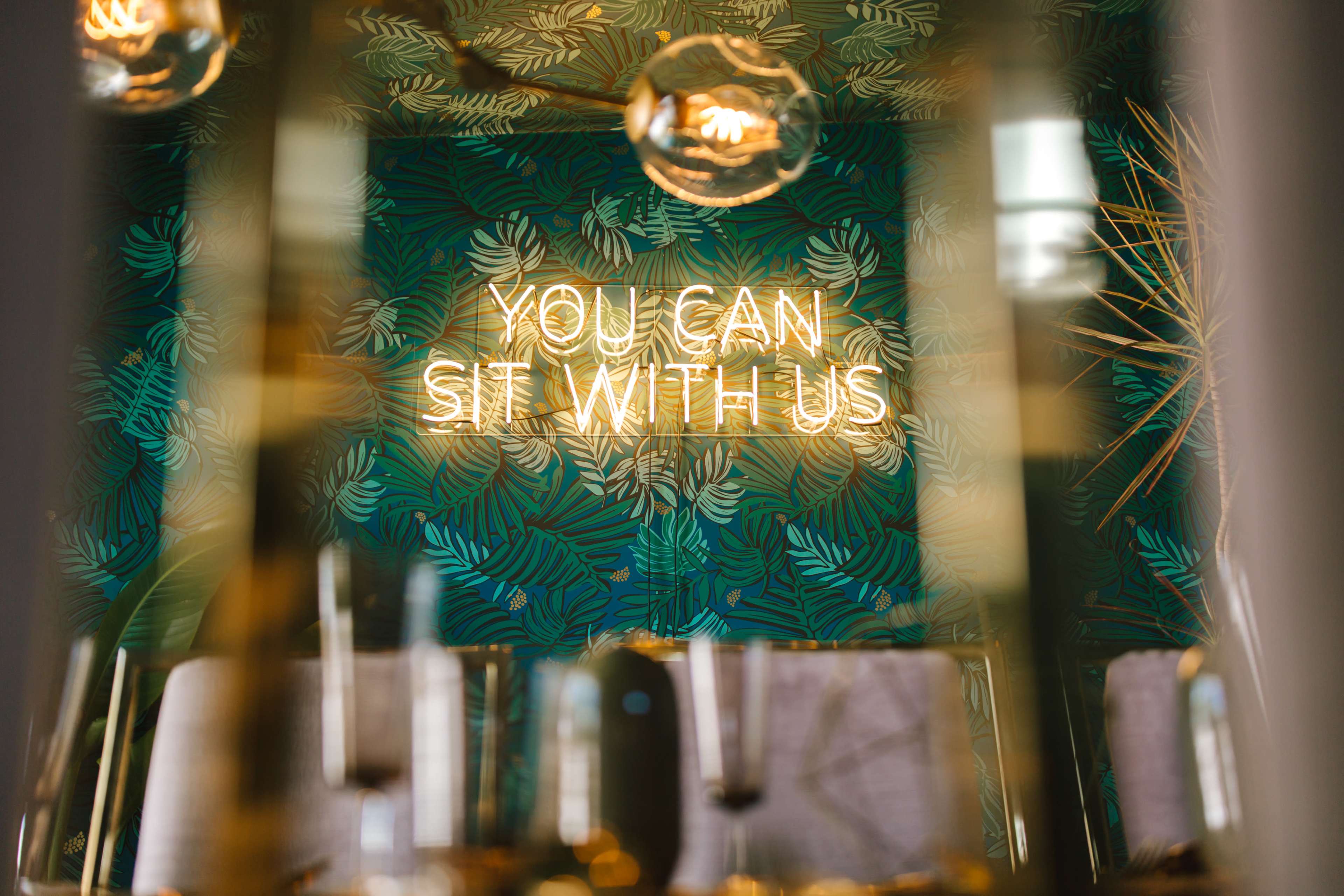 A neon sign that reads "YOU CAN SIT WITH US" against a background of tropical leaves.