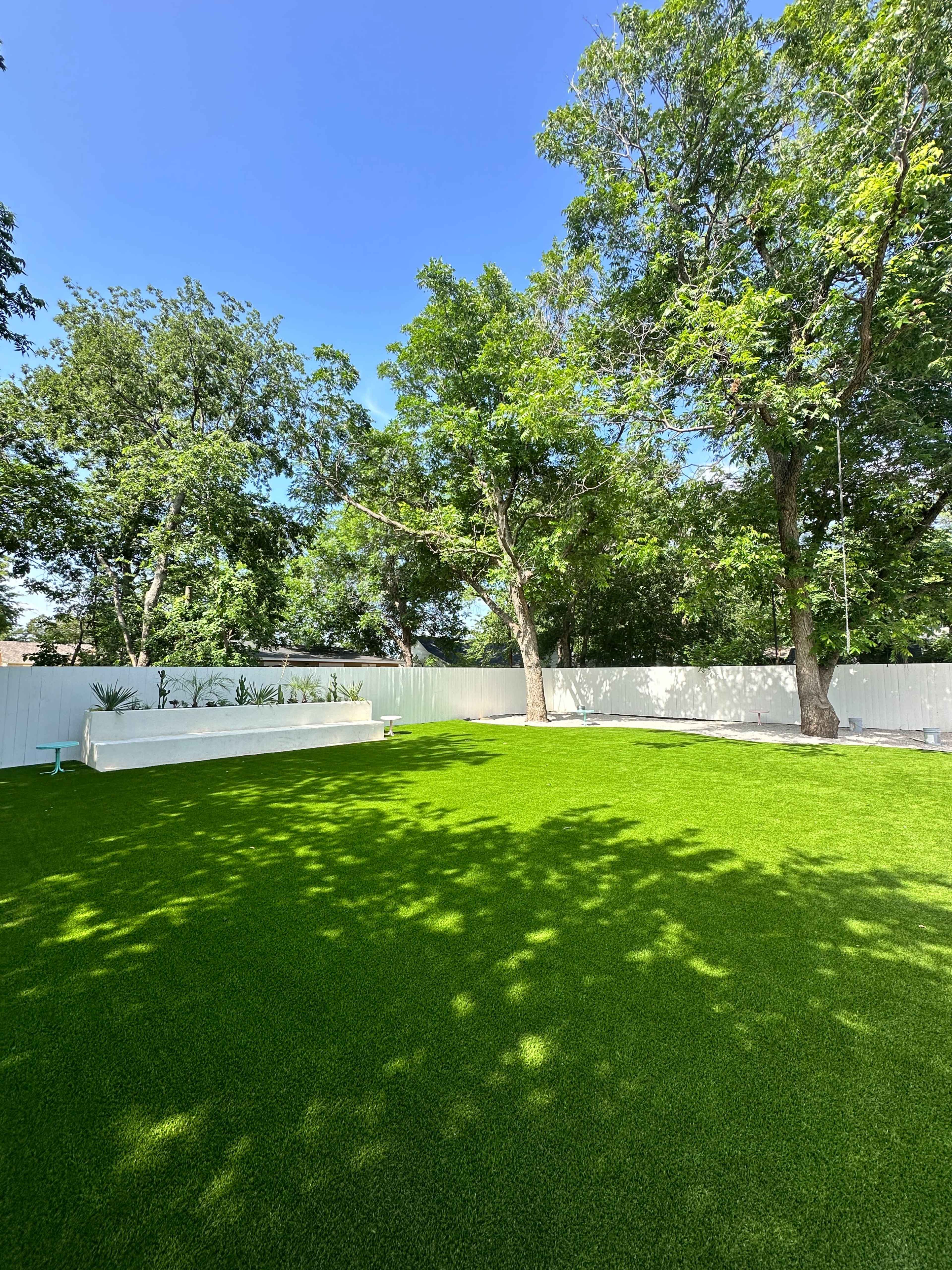 The image shows a spacious lawn with artificial turf surrounded by trees and a white fence.