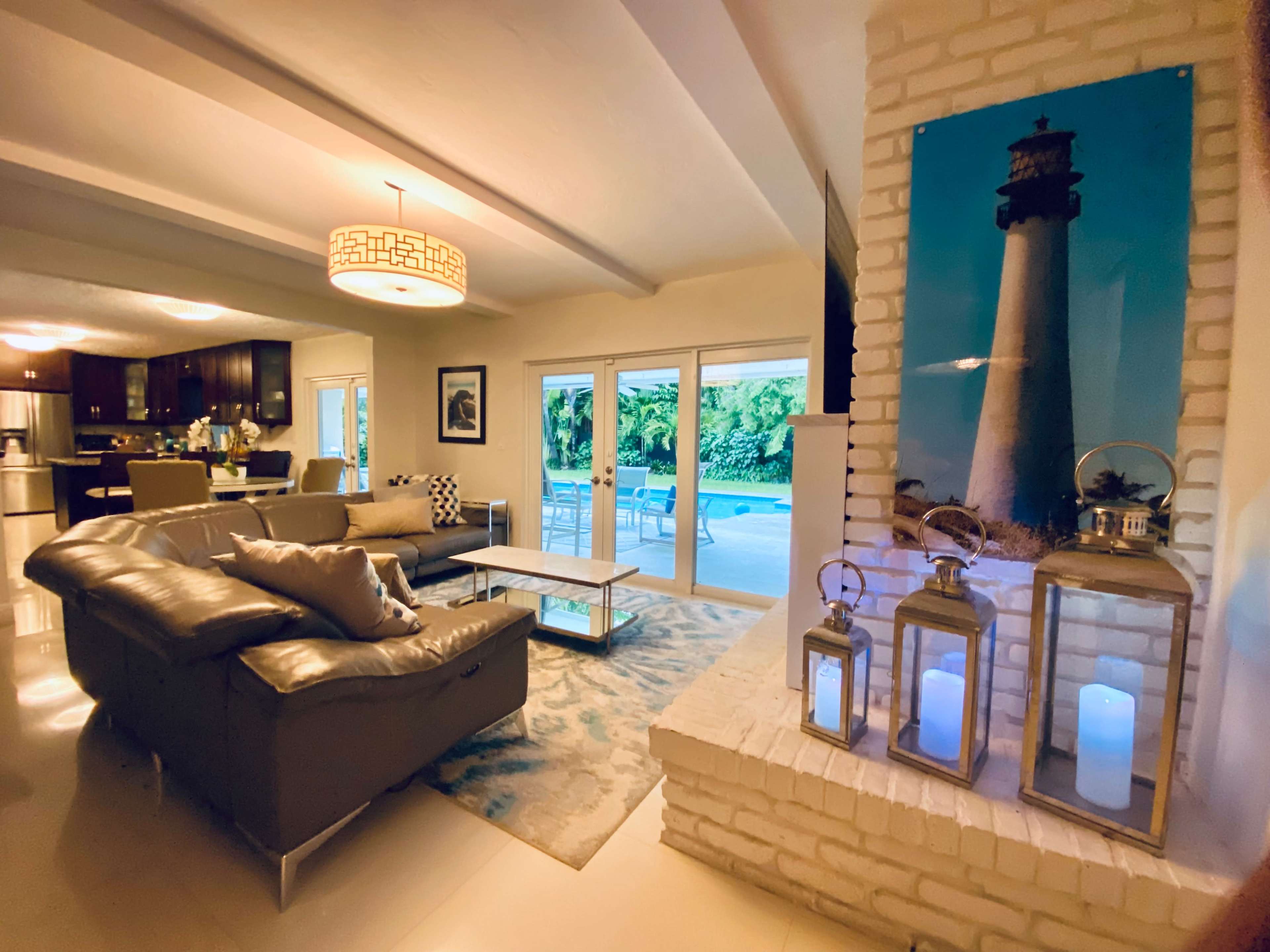 The image shows a modern living room with a gray sofa, a coffee table, and a large wall featuring a lighthouse photograph, adjacent to glass doors leading to a pool area.
