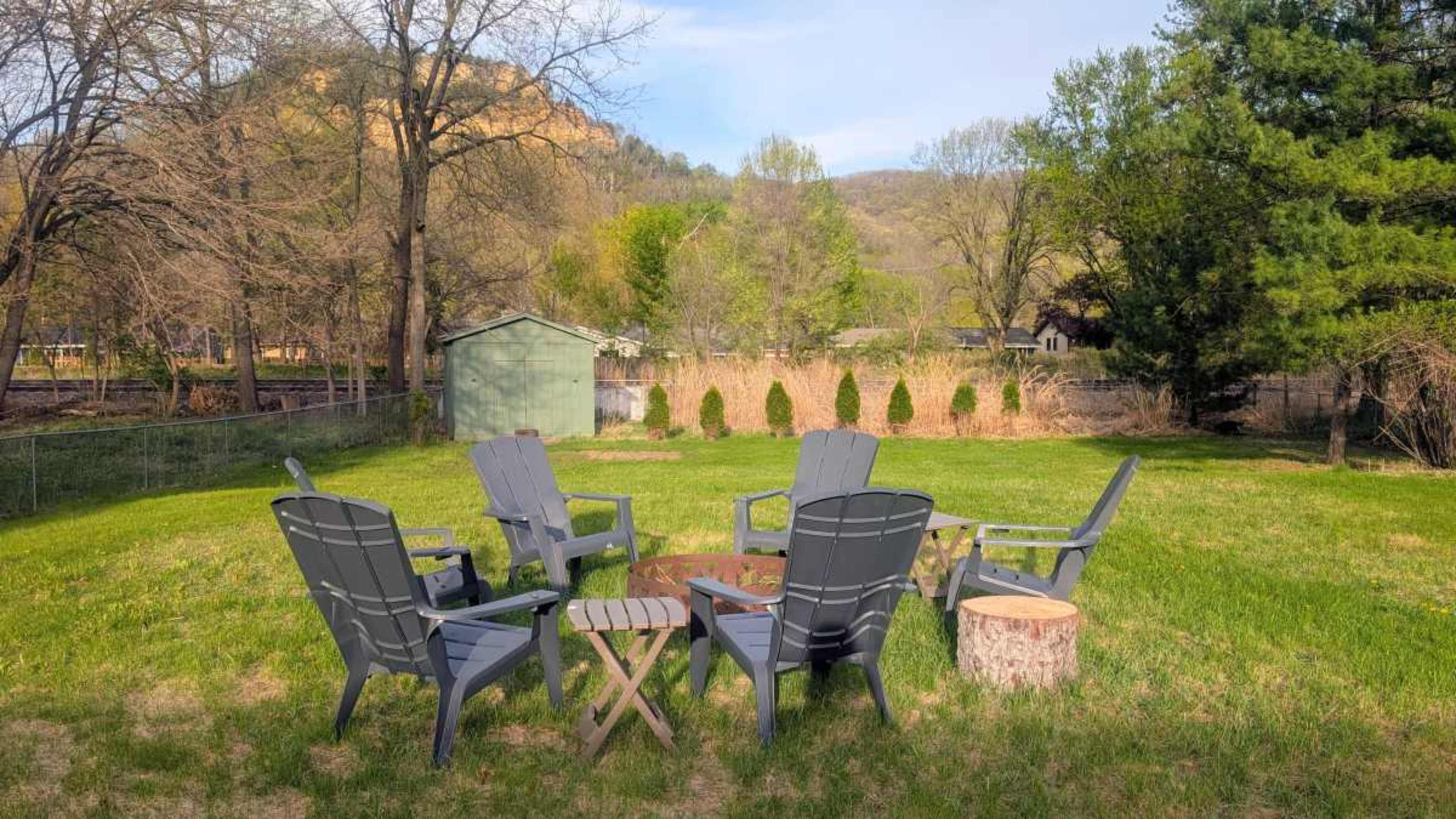 The image features a circular arrangement of six gray Adirondack chairs around a small wooden table and a fire pit in a grassy yard, surrounded by trees and hills in the background.