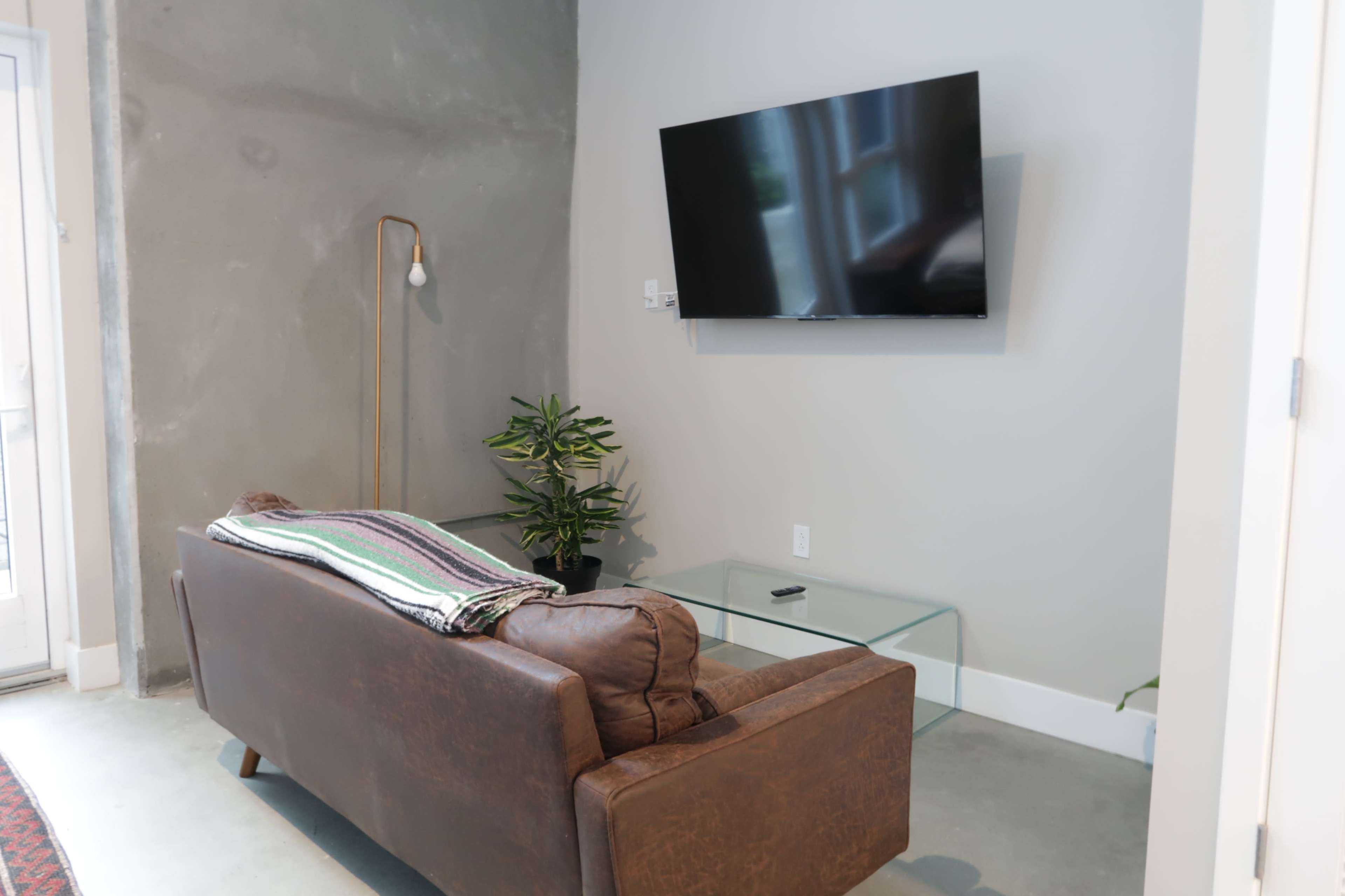 A modern living room features a brown leather sofa, a glass coffee table, a potted plant, and a wall-mounted television.