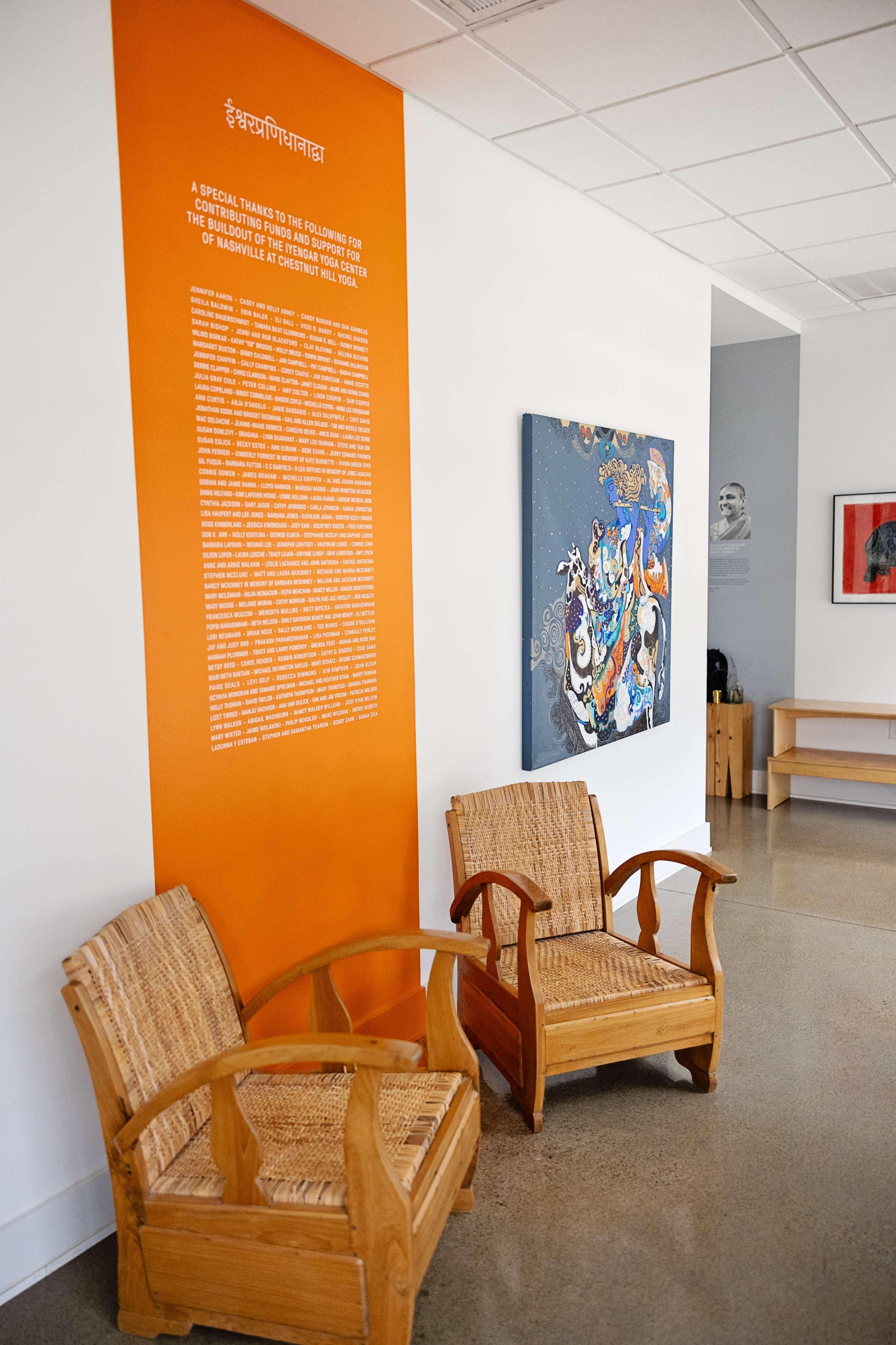 The image shows a modern interior space with two wooden chairs, an orange wall featuring text, and colorful artwork hanging nearby.