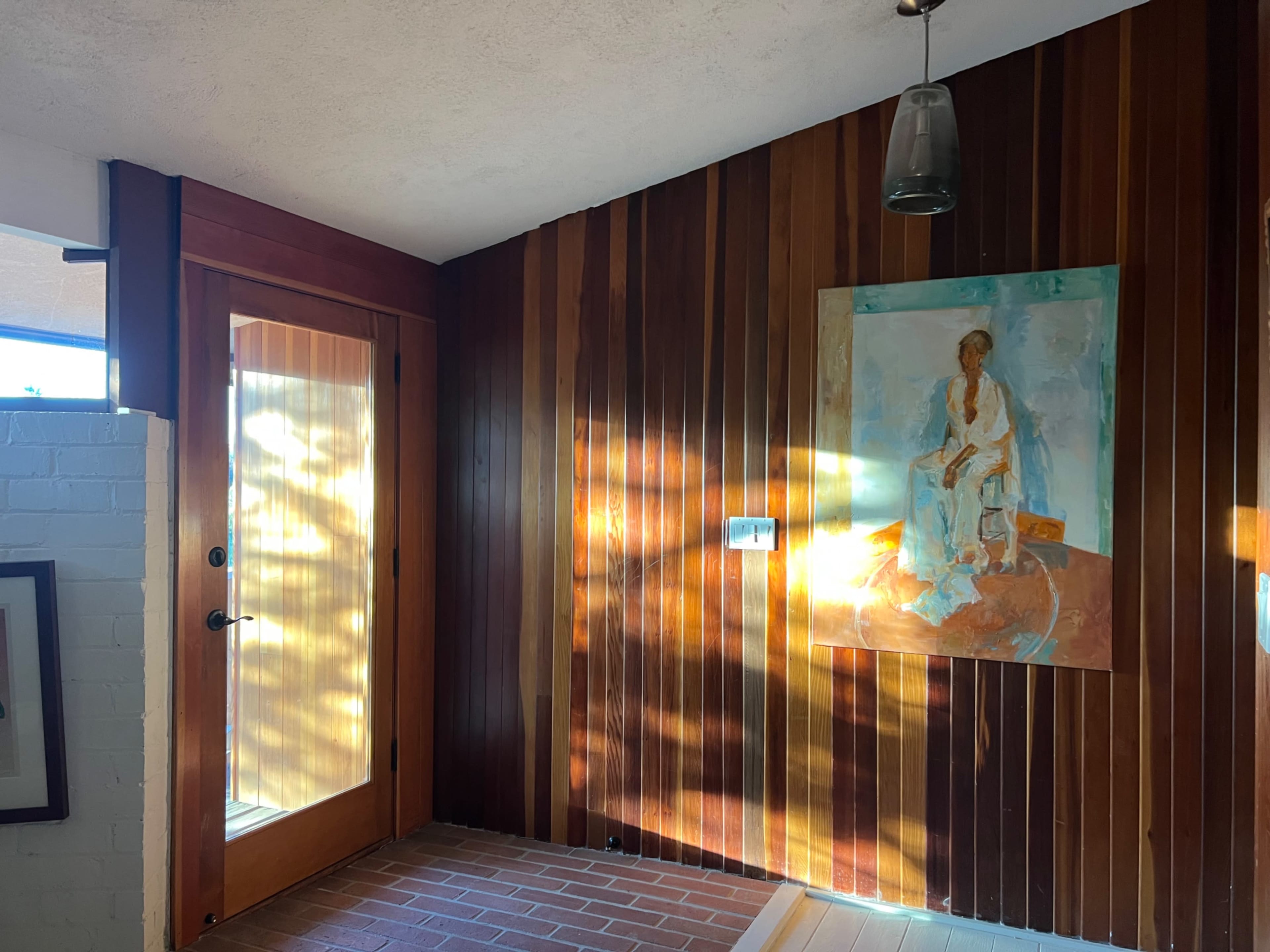 The image shows a warmly lit entryway featuring wooden walls, a glass door, and a large painting on the right.