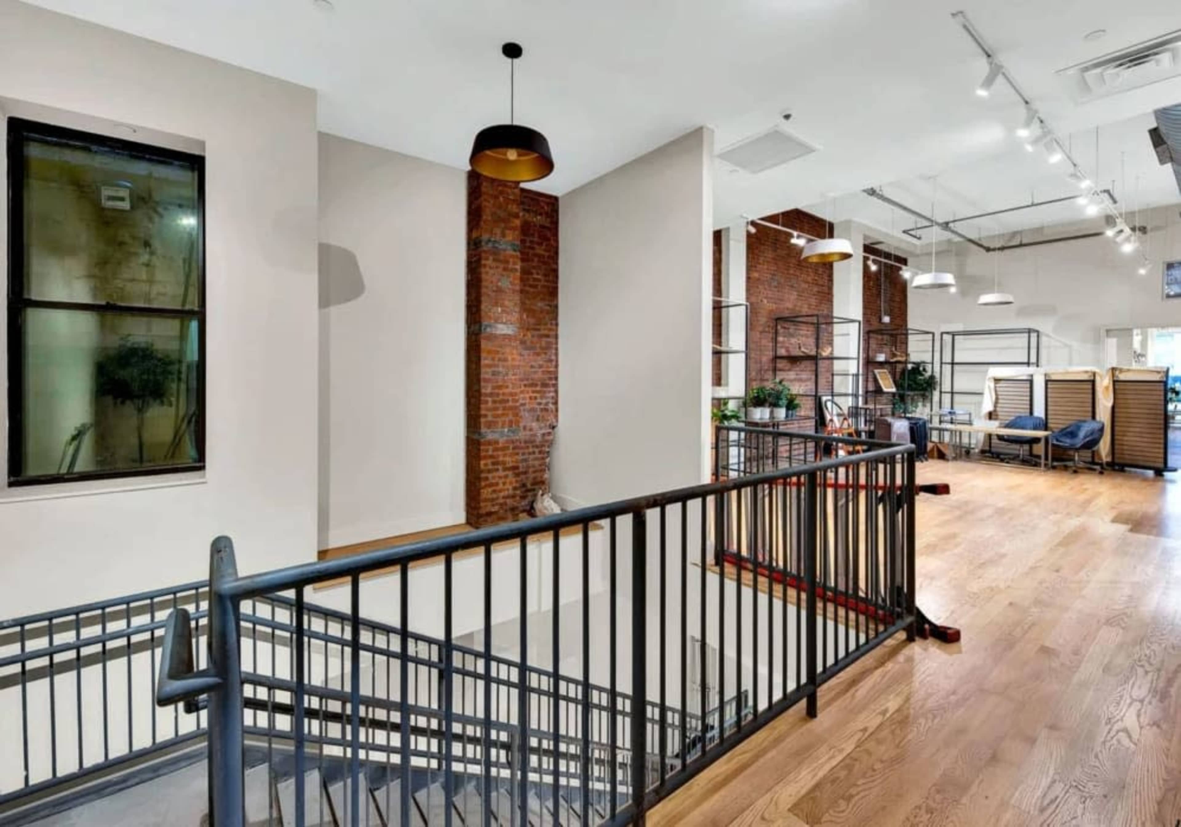 The image shows a well-lit, modern interior space featuring a staircase with metal railings, exposed brick walls, and large windows that reveal a glimpse of furniture in the background.