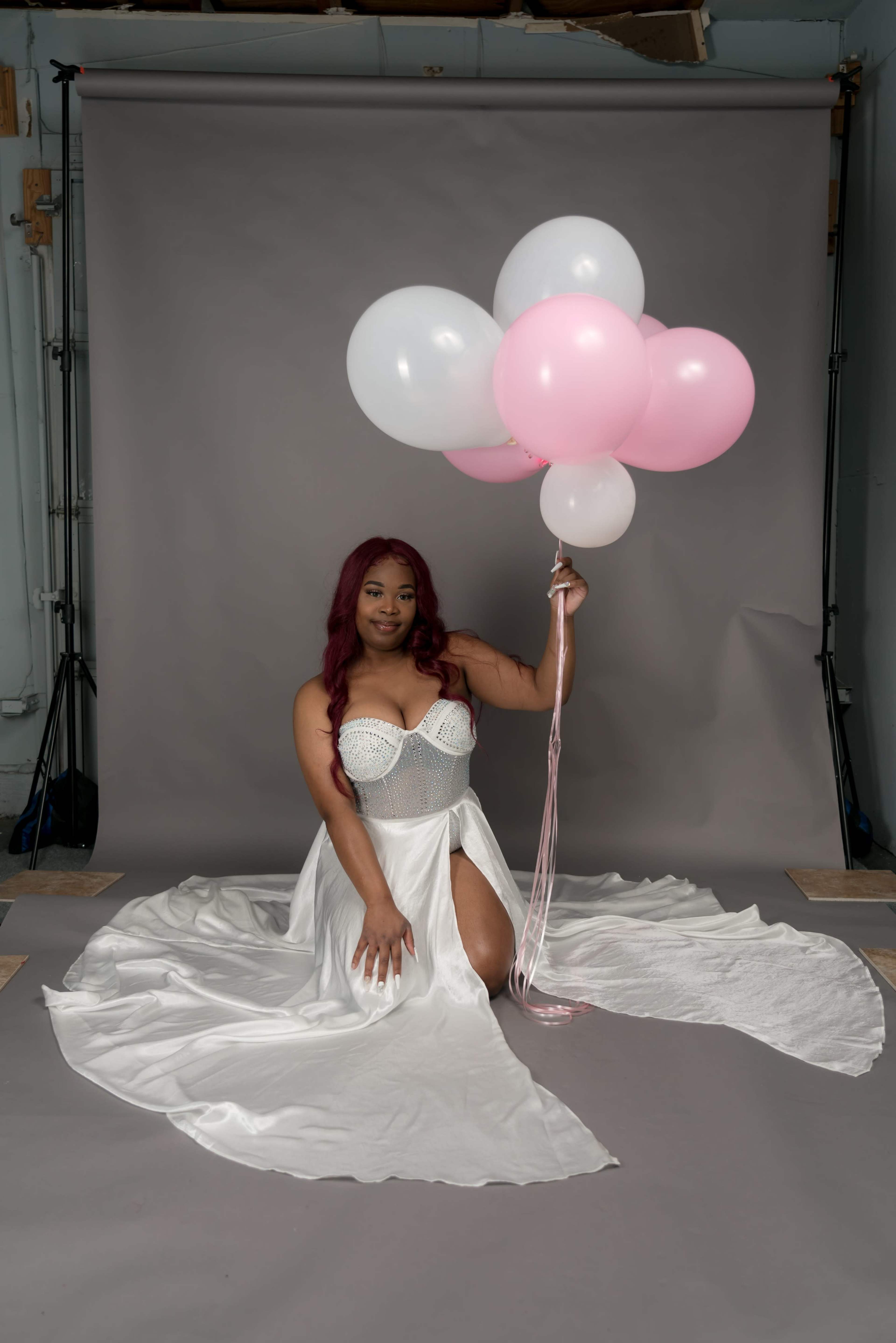 A woman in a white dress sits on a gray floor, holding a cluster of pink and white balloons.