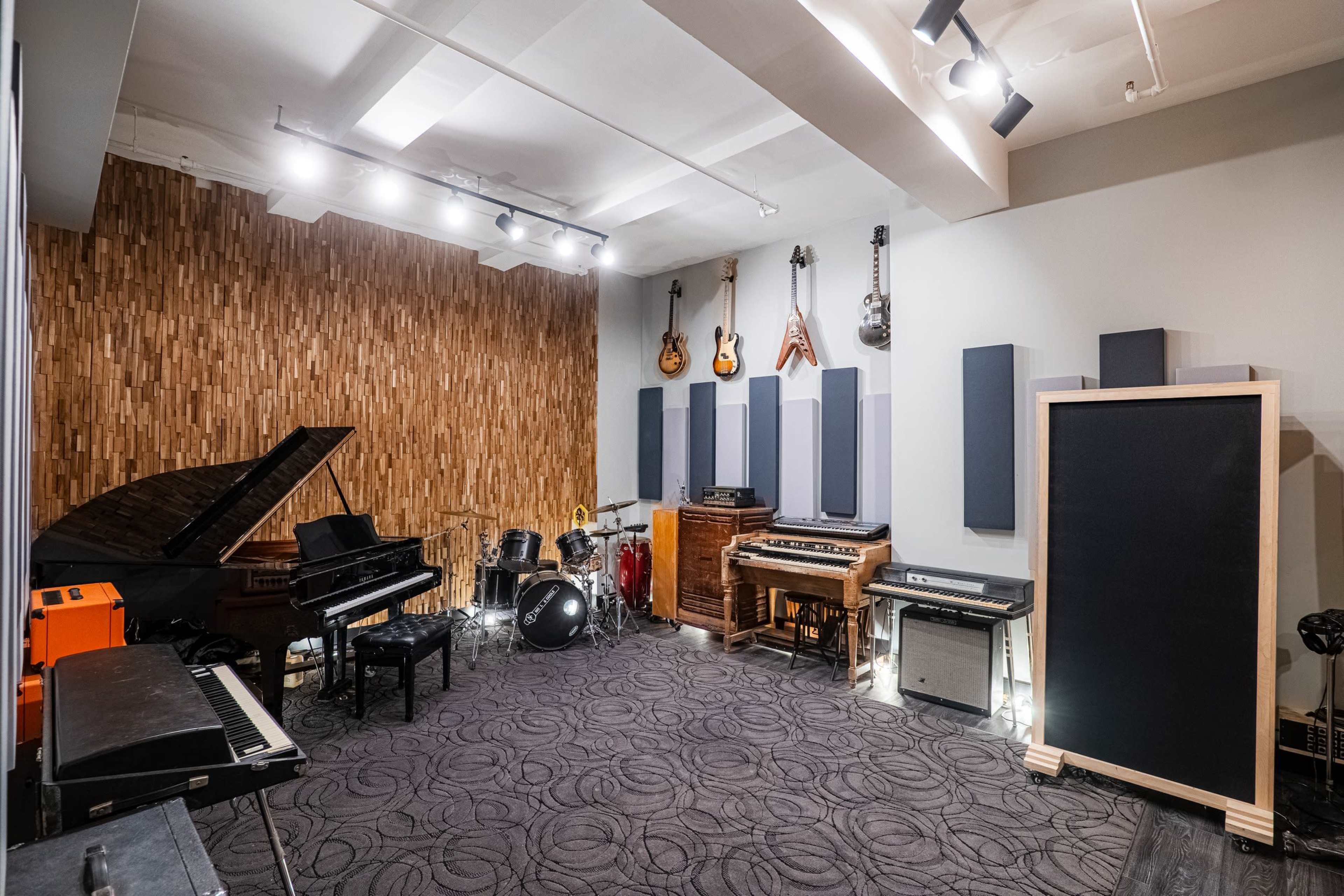 The image shows a music studio featuring various musical instruments, including a grand piano, drum set, and electric keyboards, arranged along wooden and soundproof walls.