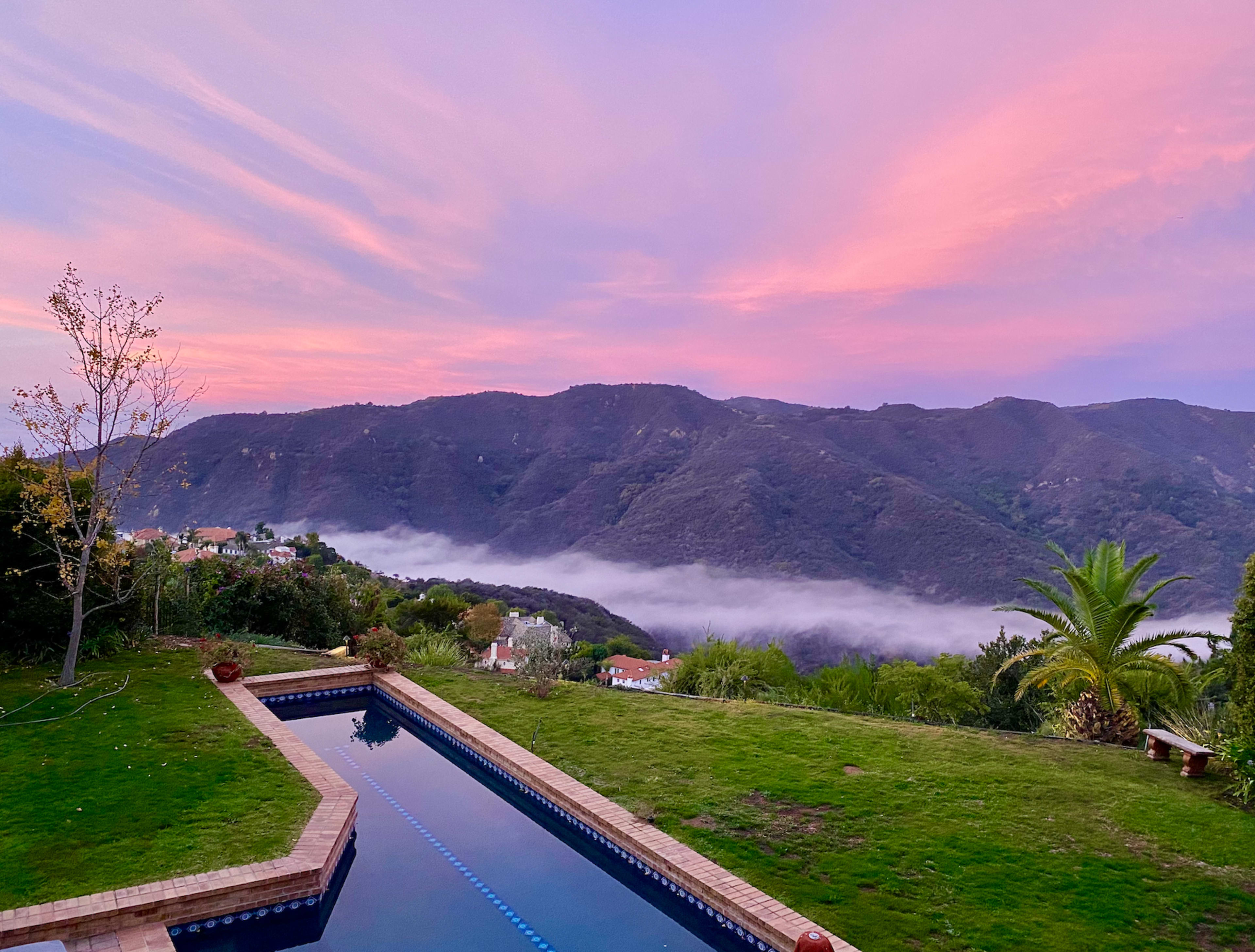 A swimming pool bordered by grass and plants, overlooking a mountainous landscape with fog and a colorful sky during sunset.