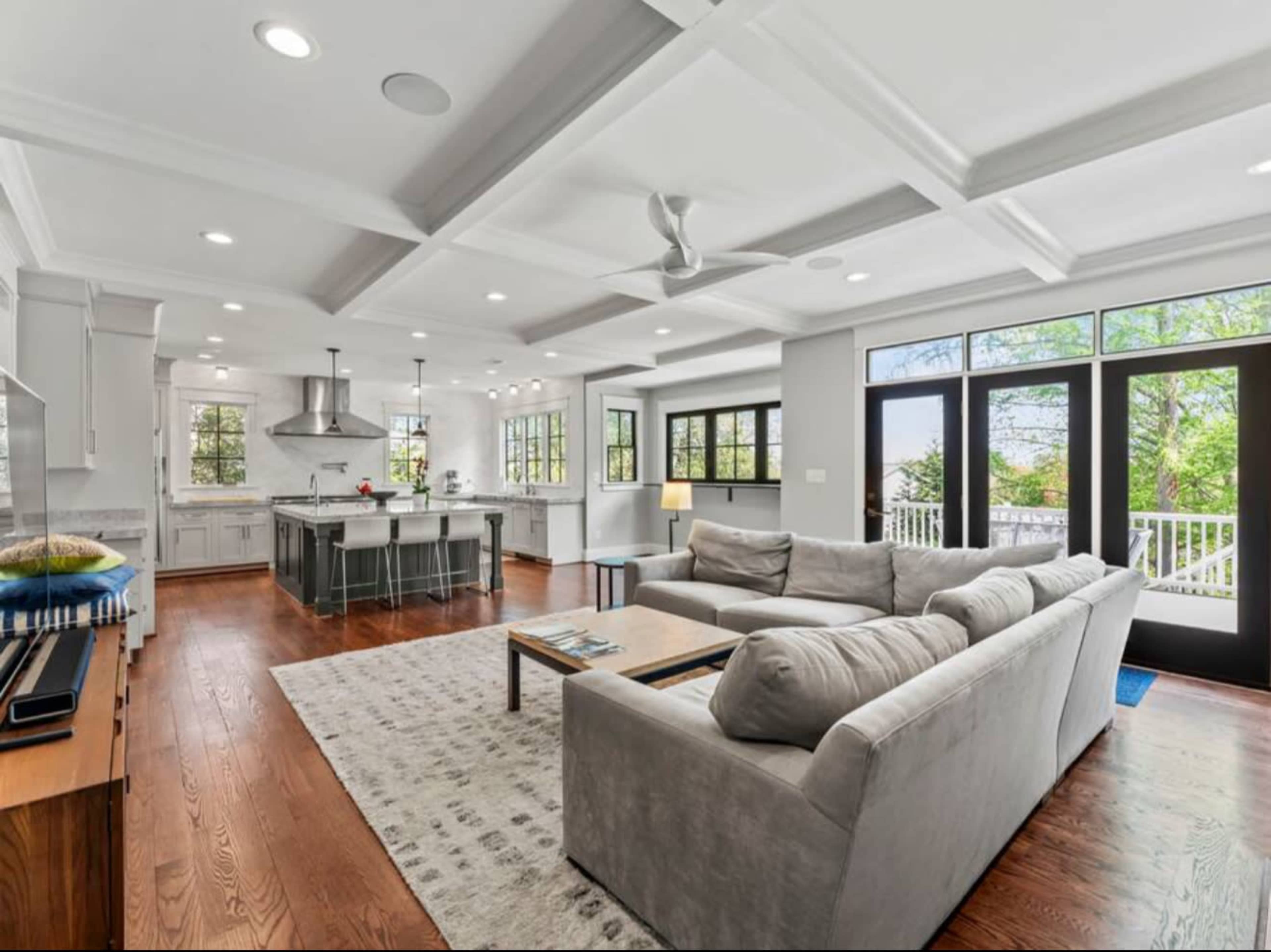 The image shows a spacious living area with a gray sectional sofa, a wooden coffee table, and large windows allowing natural light to enter, adjoining a modern kitchen equipped with stainless steel appliances.