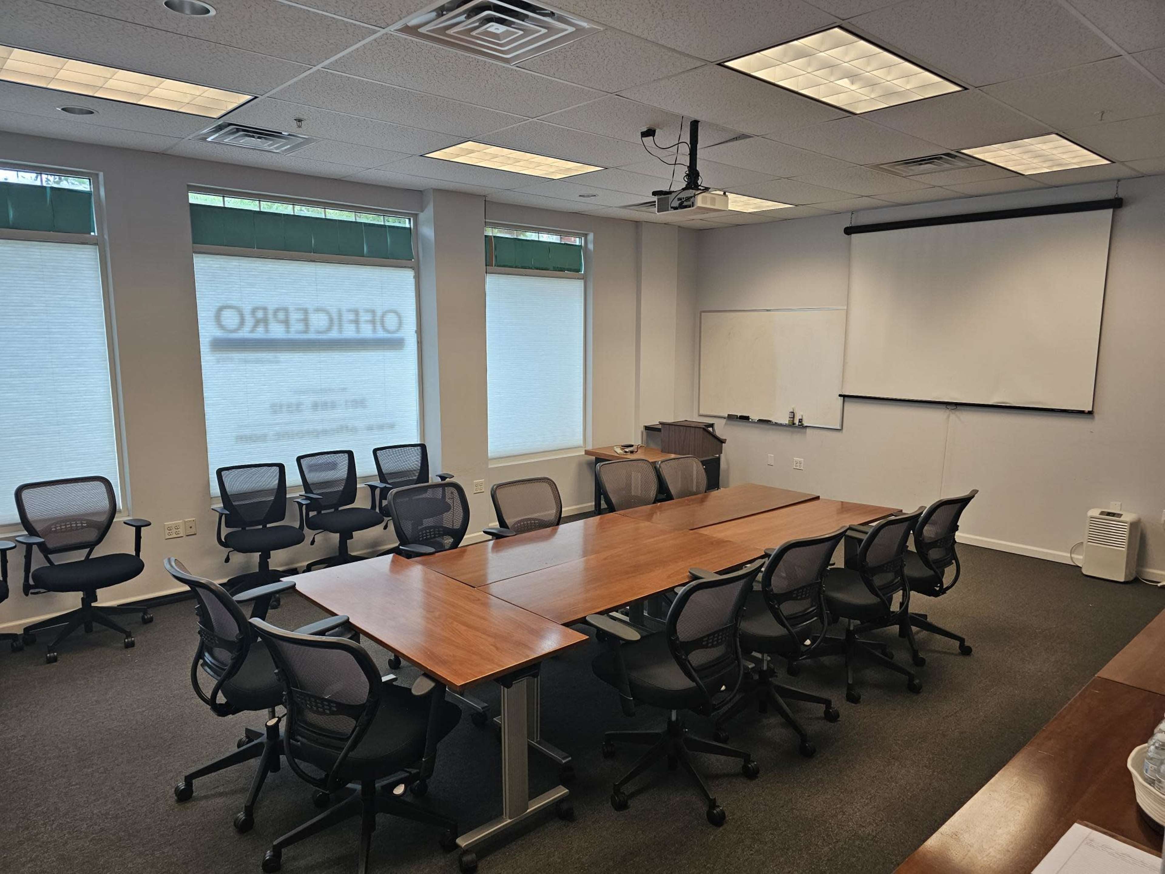 A conference room with a long wooden table surrounded by chairs, a projector mounted on the ceiling, and windows with blinds.