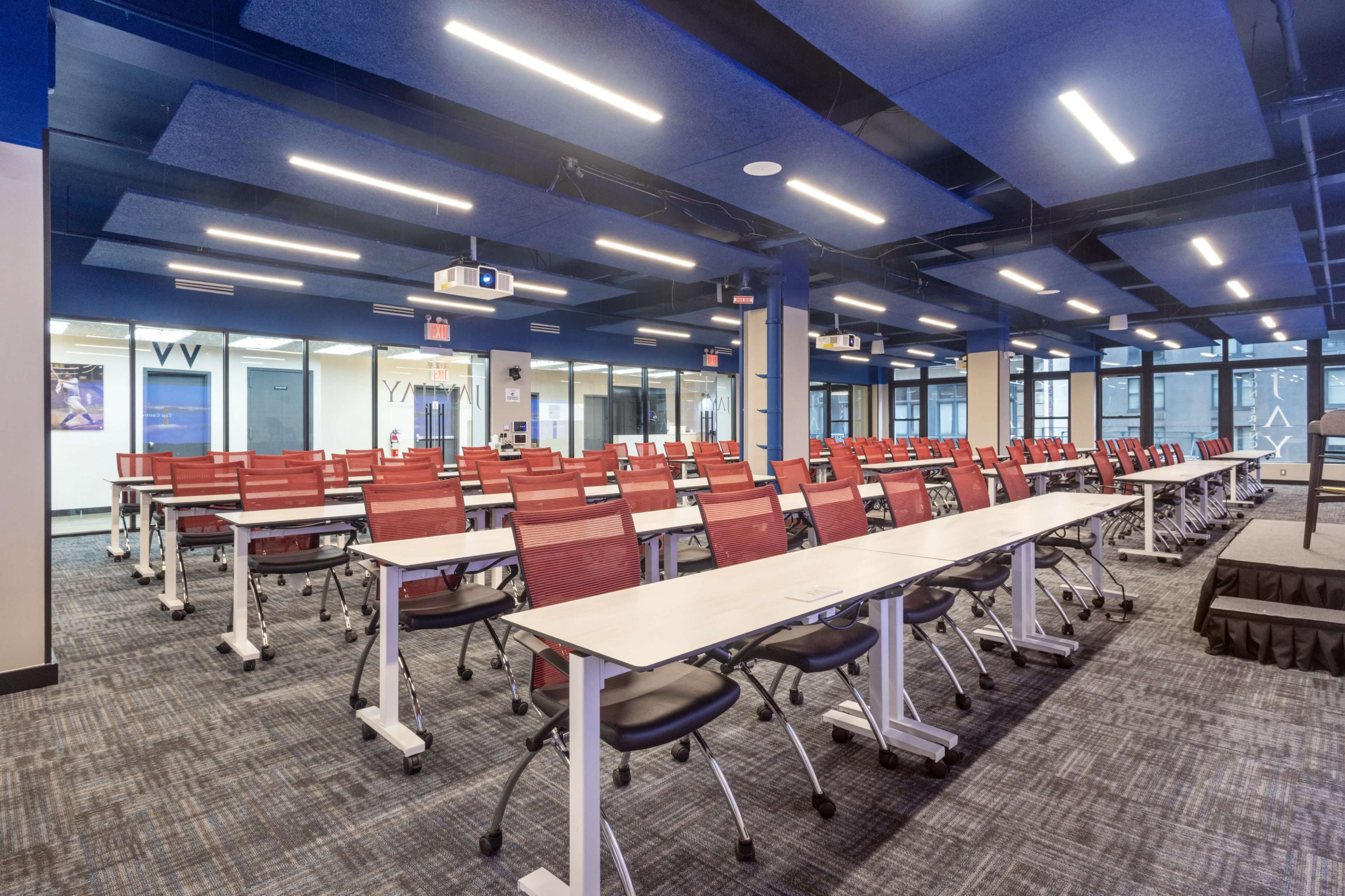 A spacious modern classroom with rows of red chairs and white tables, equipped with overhead lighting and a projector.