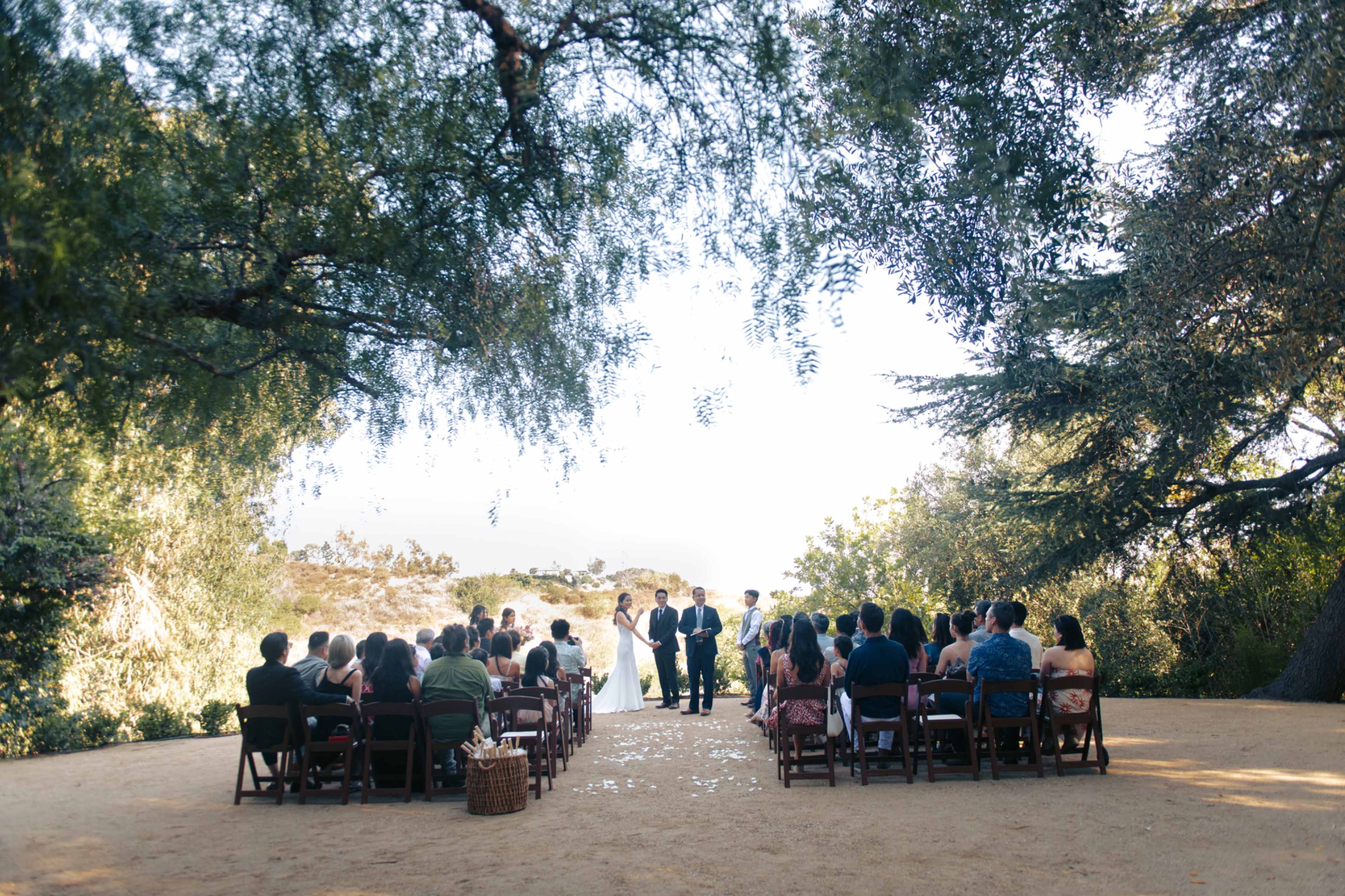 A couple stands in front of their guests during an outdoor wedding ceremony surrounded by trees.