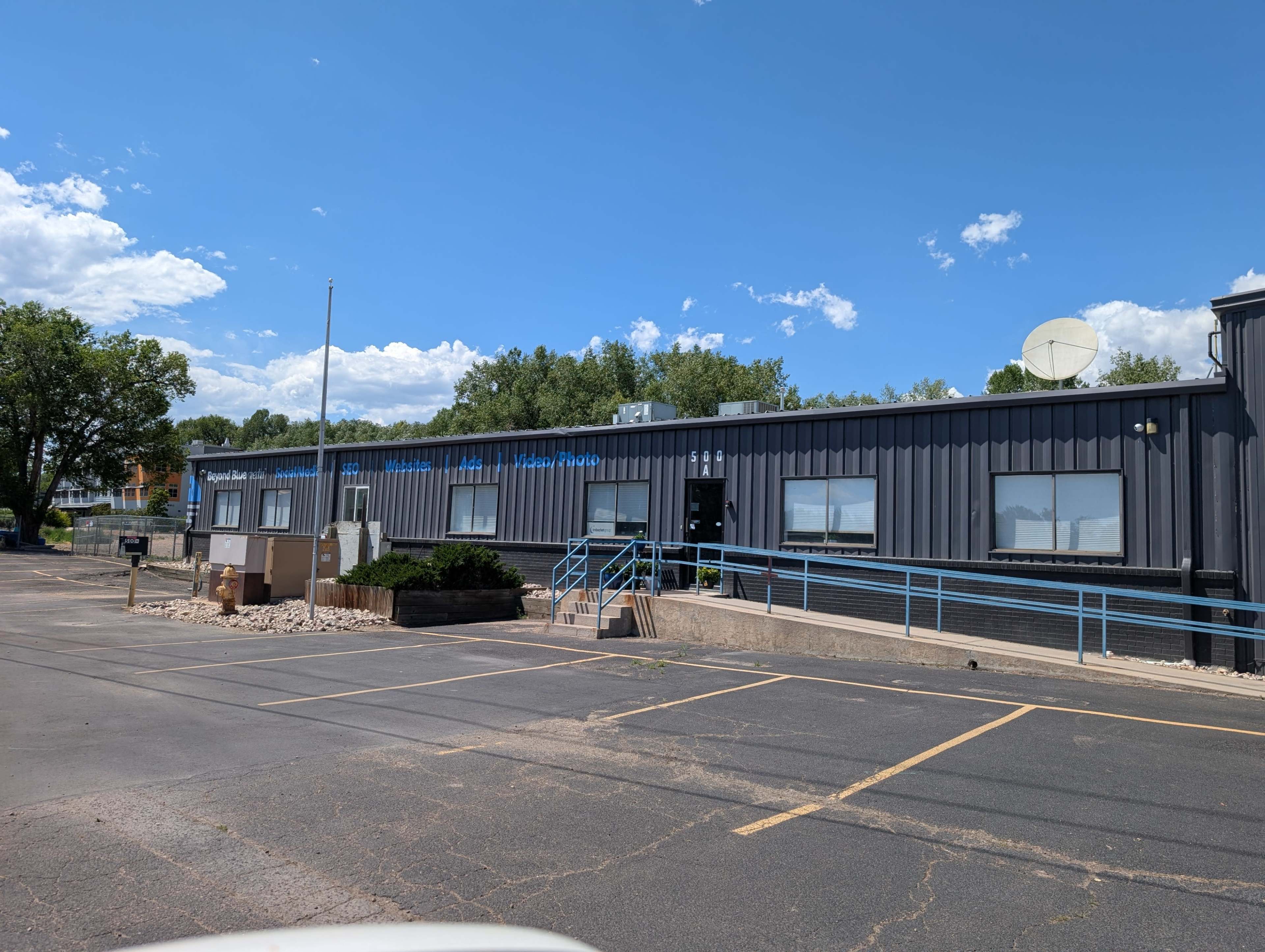 The image shows a gray metal building with windows, a blue railing, and a flagpole surrounded by a paved parking area and some landscaping.