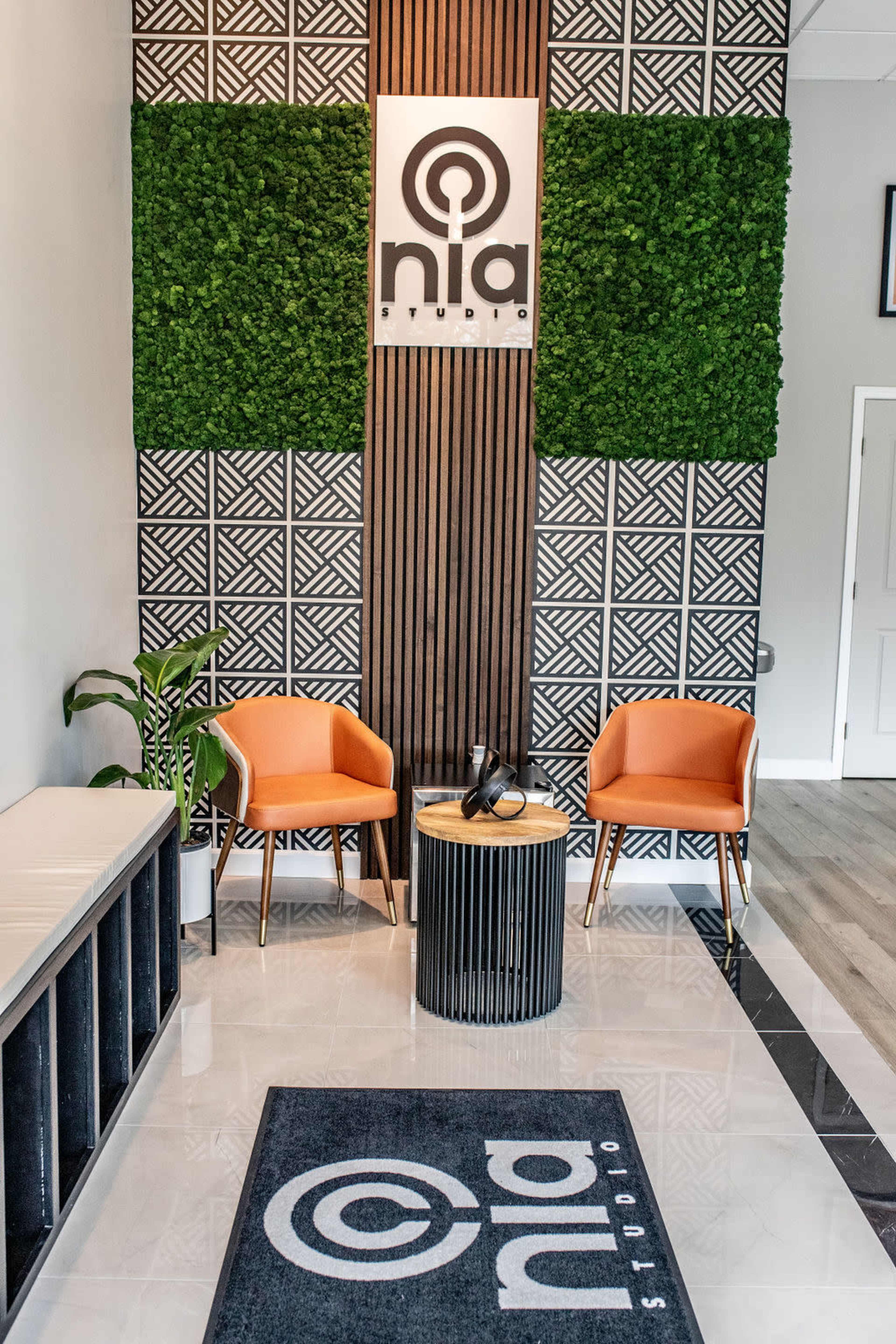 A modern reception area featuring two orange chairs, a circular table, and a green plant against a patterned wall.