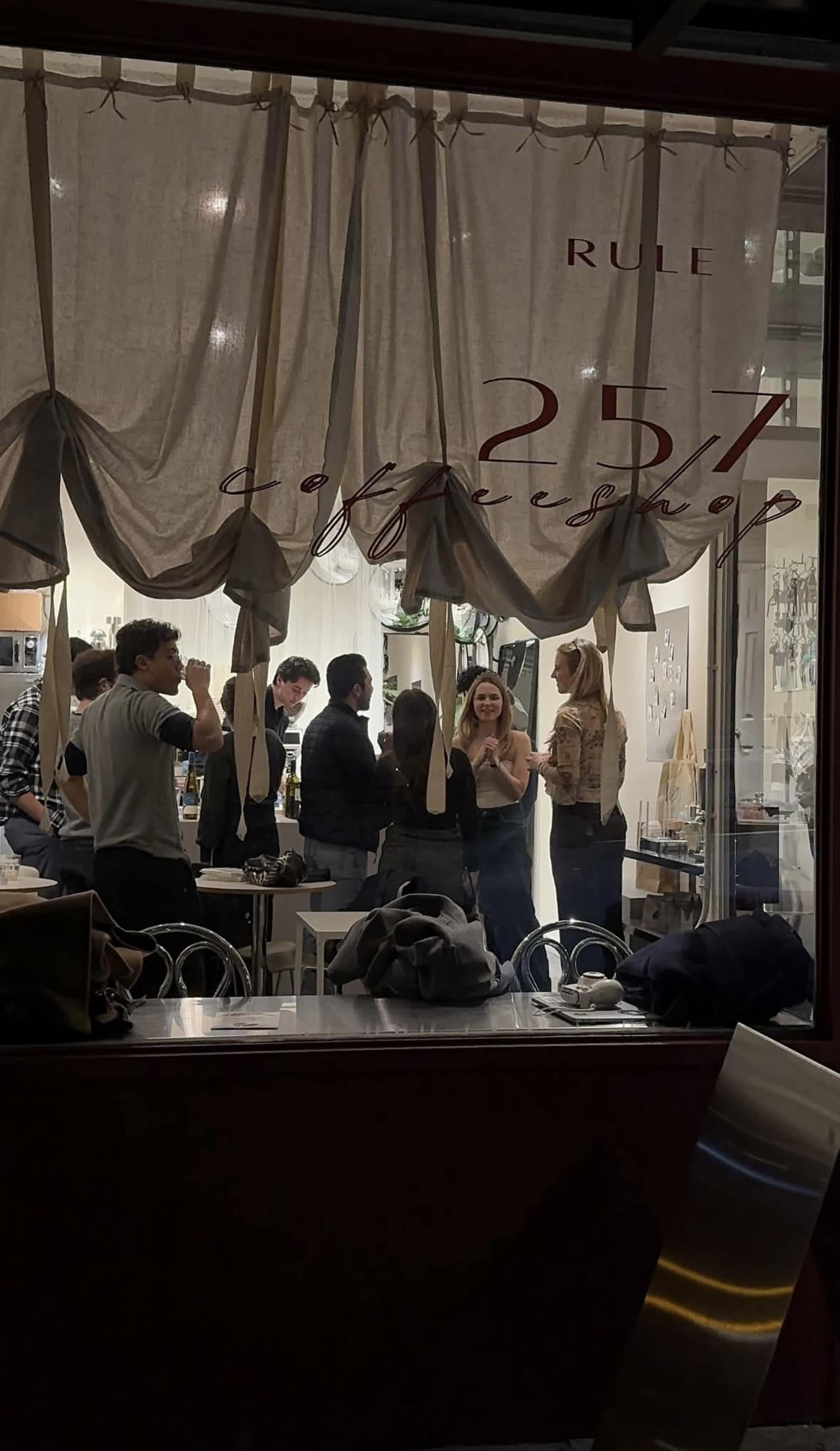A group of people is gathered inside a café, seen through a large window with fabric drapes above.