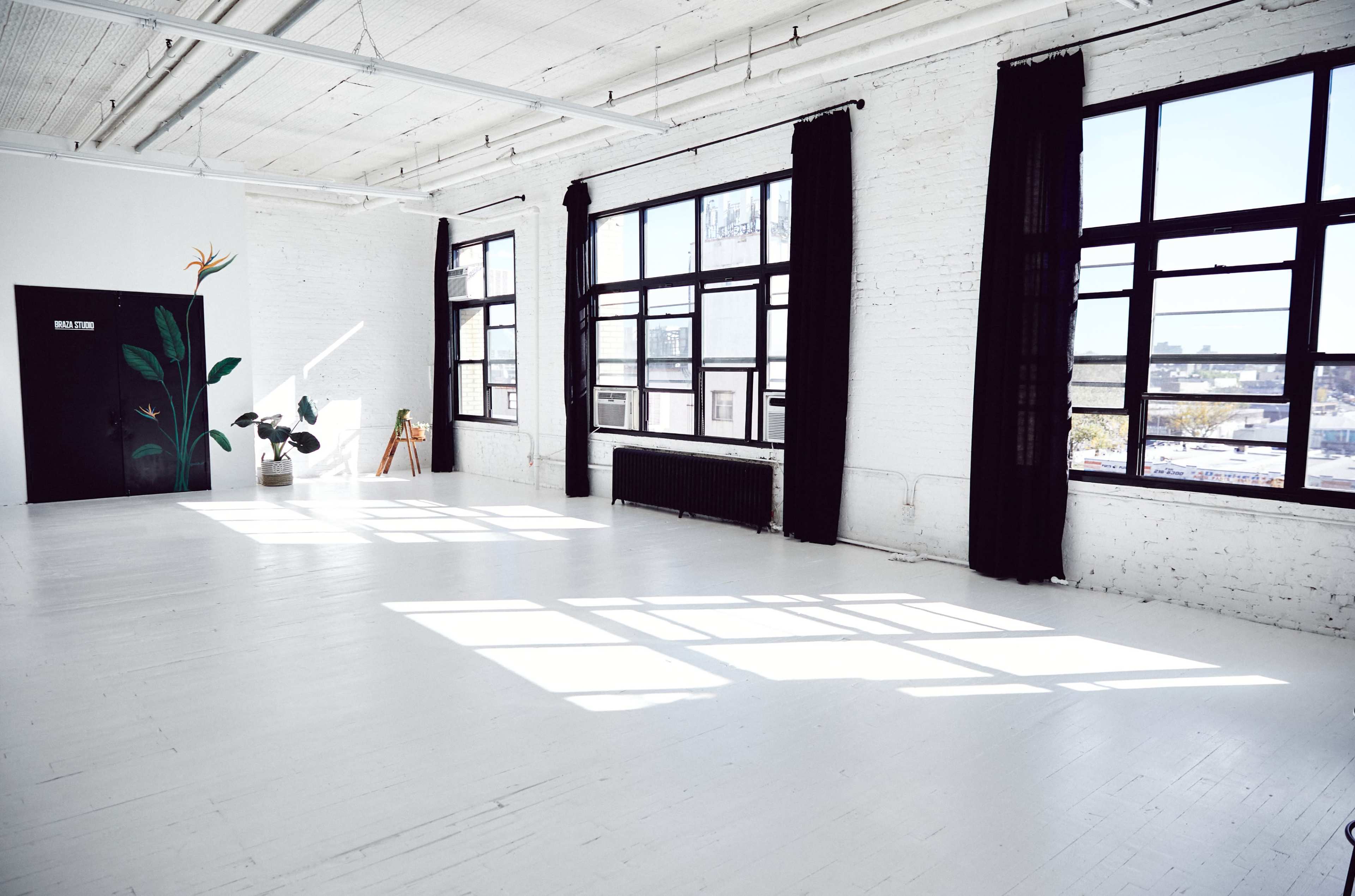 A bright, airy studio space with large windows, whitewashed walls, and a minimalistic design featuring a potted plant and an easel.