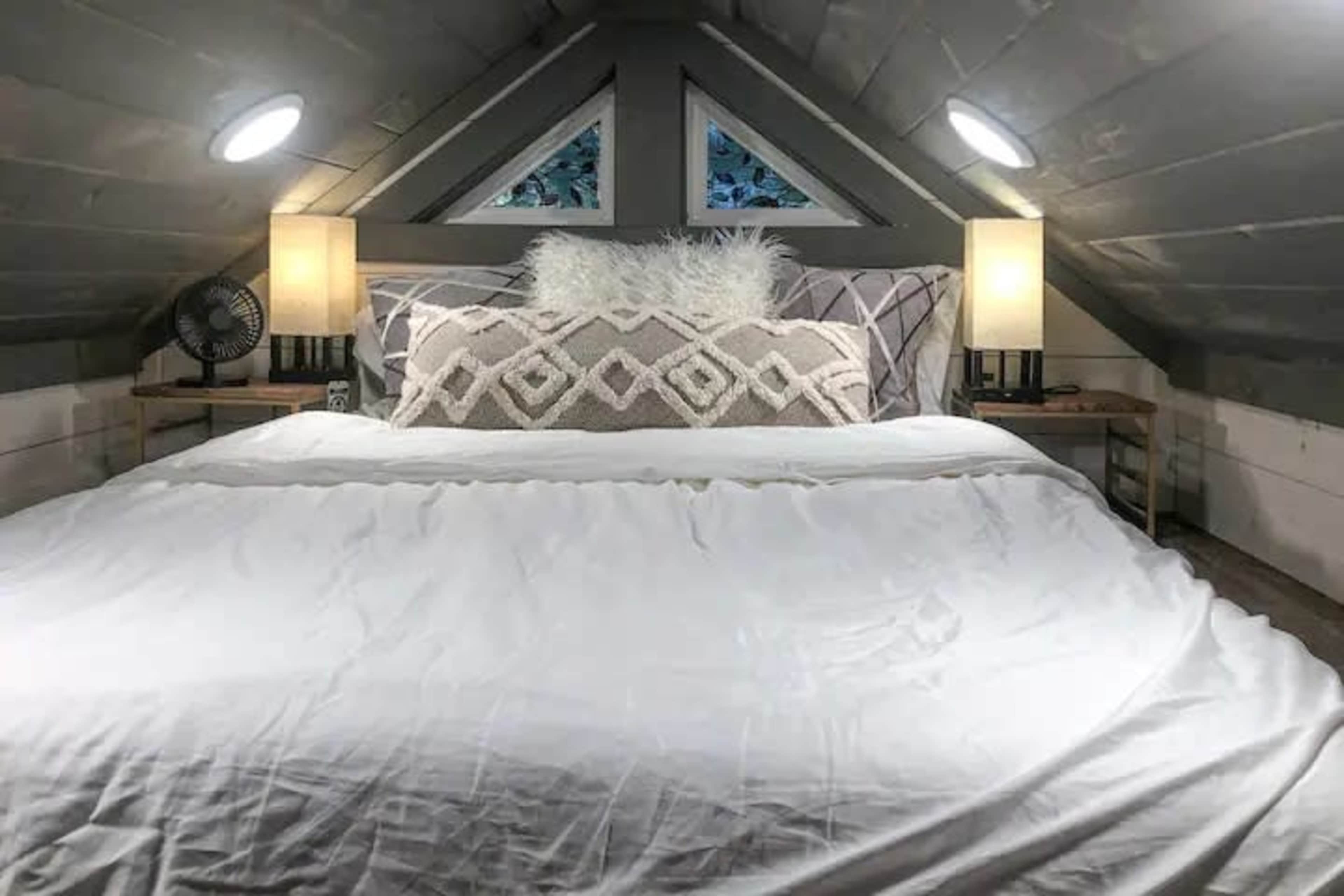 A cozy attic bedroom featuring a large bed with a white bedspread, two bedside lamps, and decorative pillows, flanked by angled wooden walls and a ceiling with triangular windows.