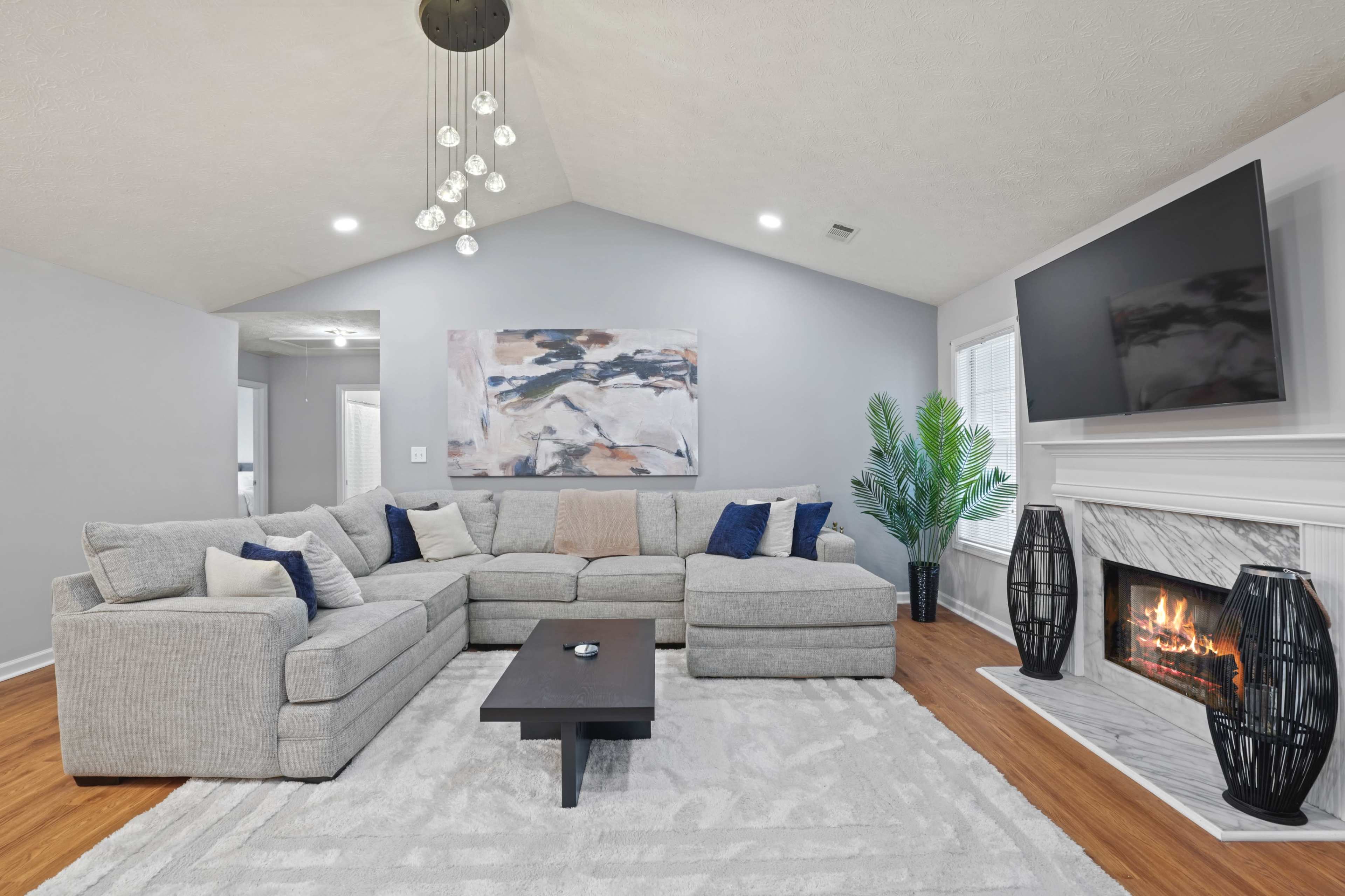 A modern living room features a large sectional sofa, a low coffee table, a TV mounted above a fireplace, and a decorative painting on the wall.