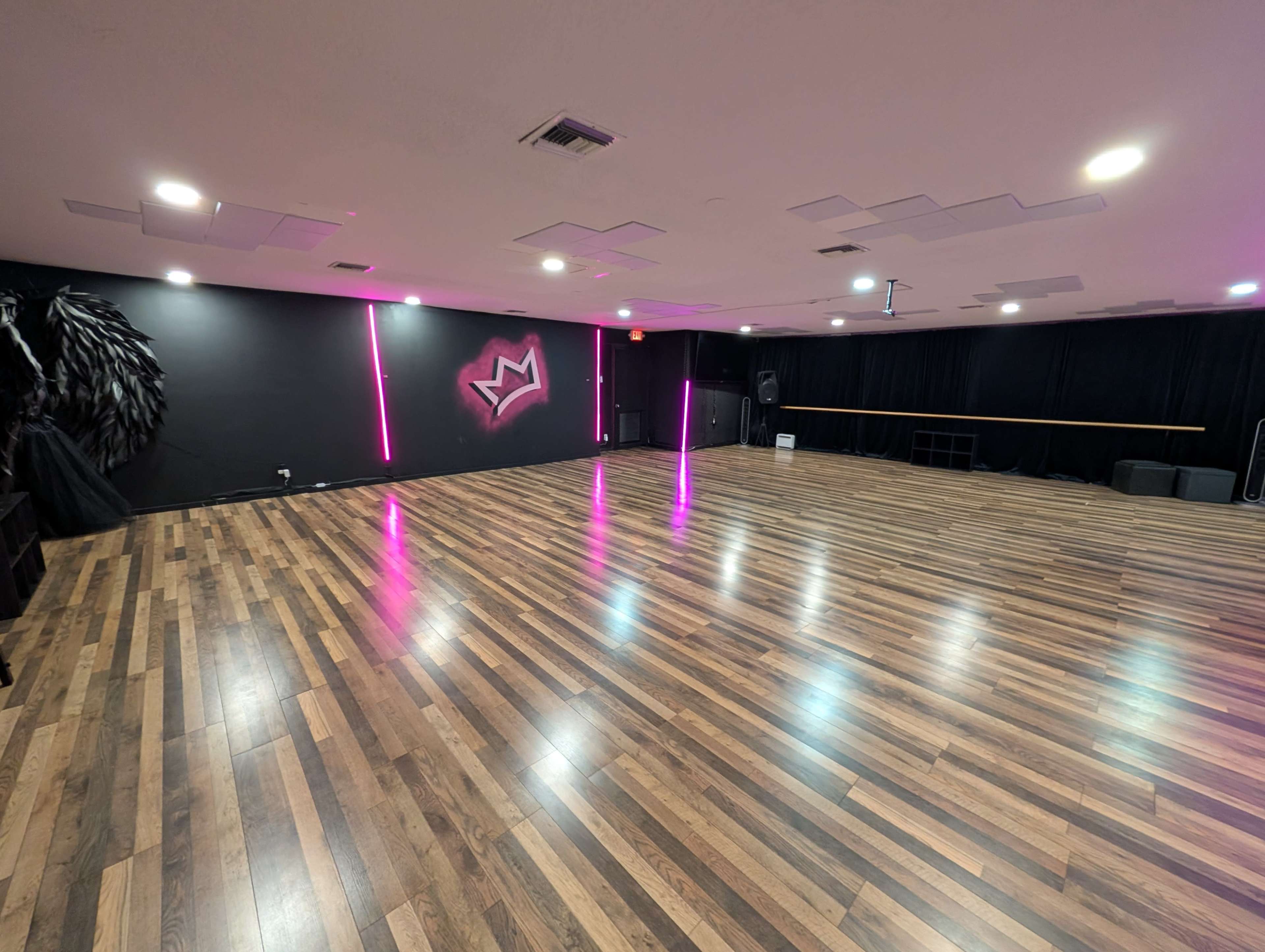 The image shows a spacious dance studio with wooden flooring, black walls, and two neon pink light fixtures.