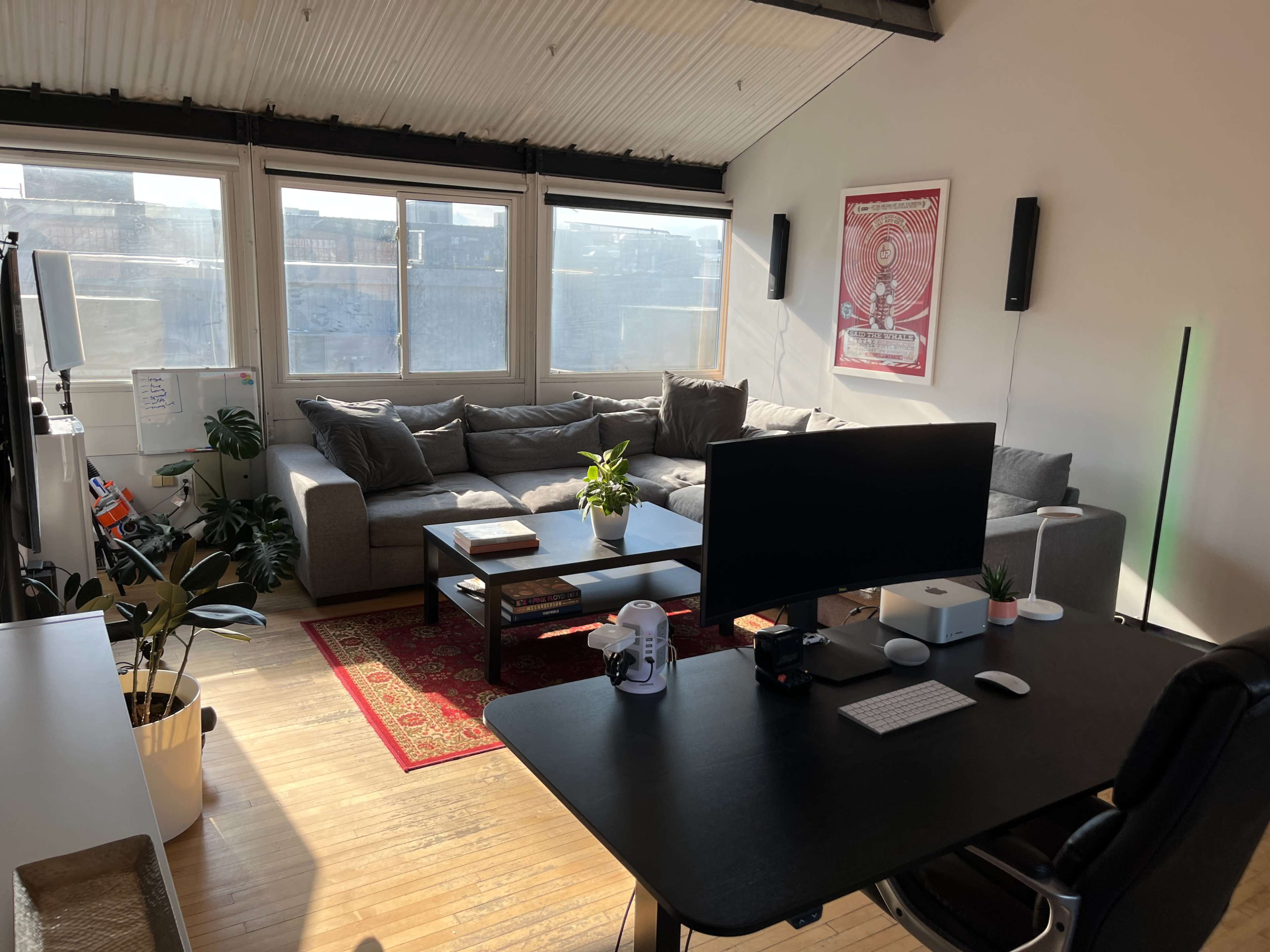 A cozy living room with a large gray sectional sofa, a coffee table, a desk with a computer, and plants arranged throughout the space.