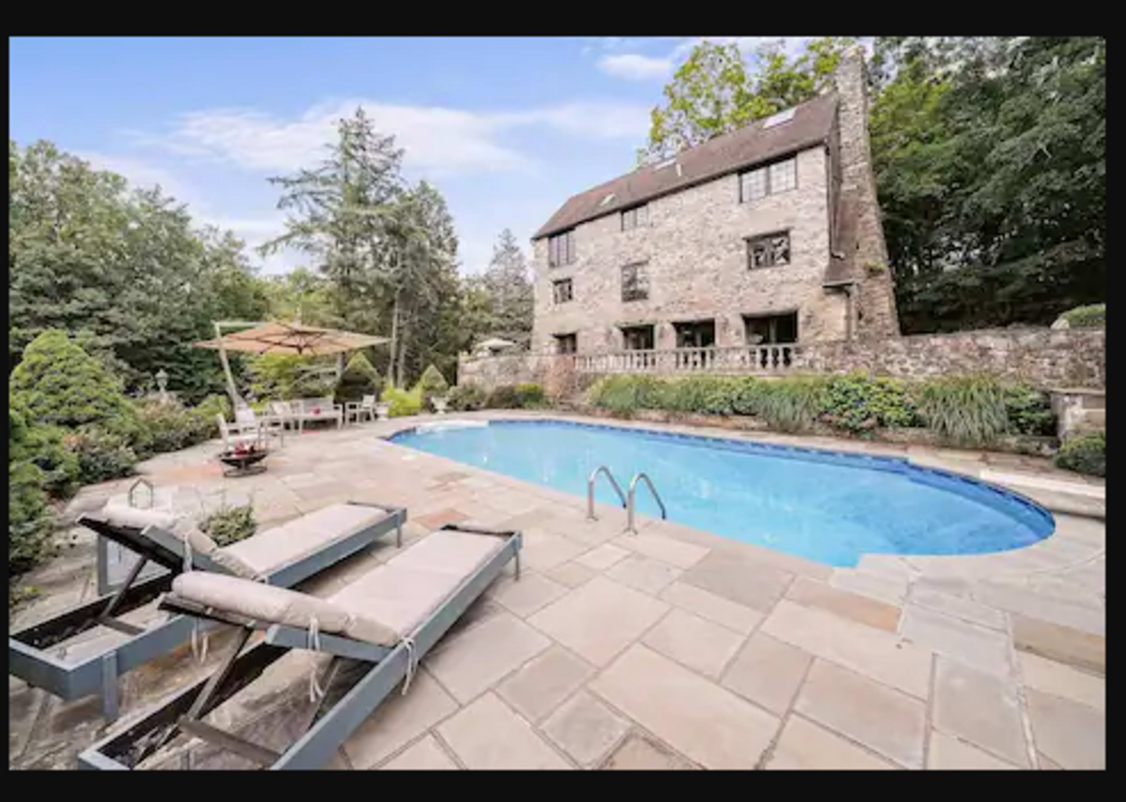 A stone house with a swimming pool surrounded by a stone patio, lounge chairs, and lush greenery.