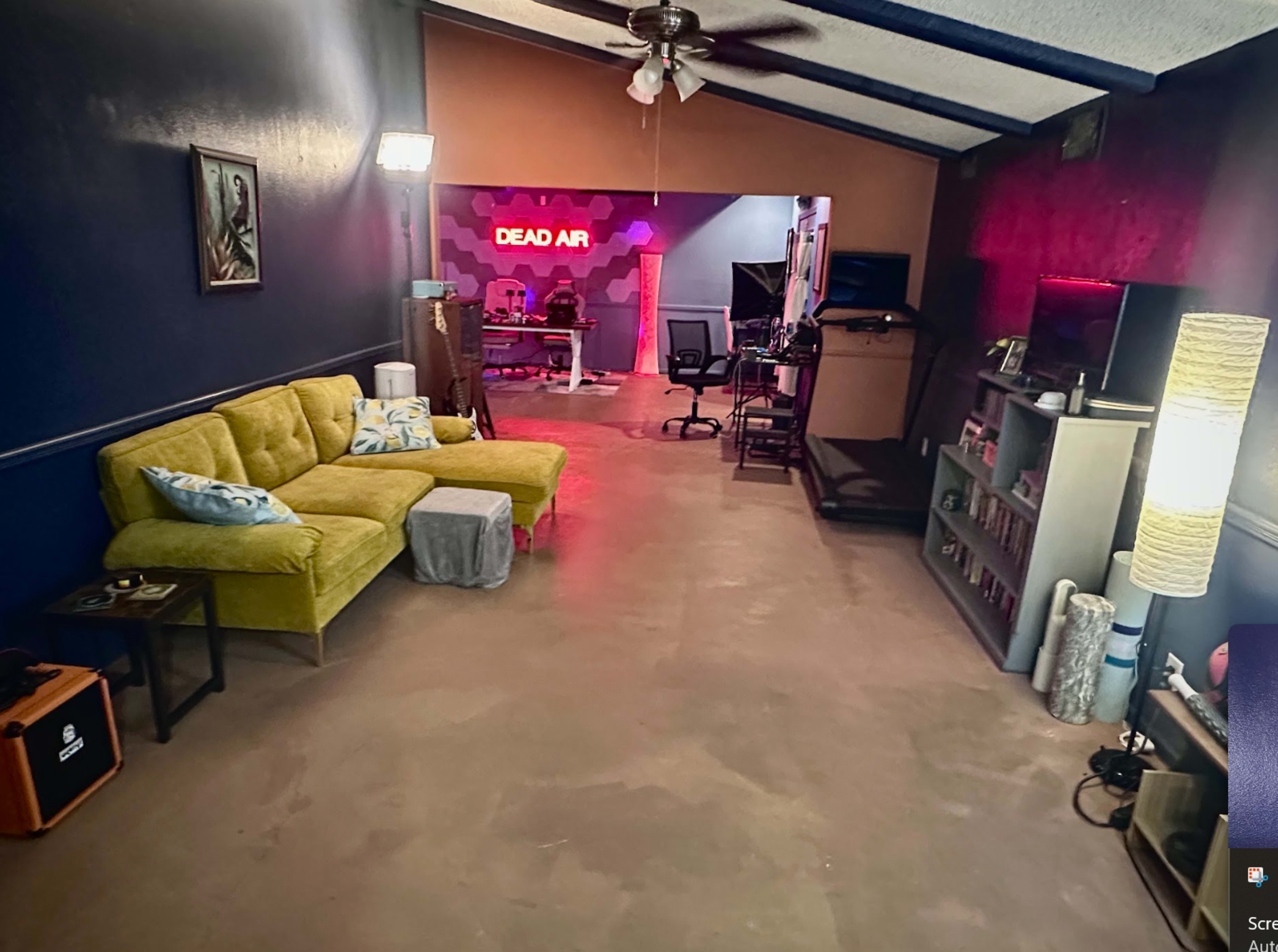 The image shows a spacious room featuring a yellow couch, a small table, a bookshelf, and a studio area with illuminated signs and various equipment.