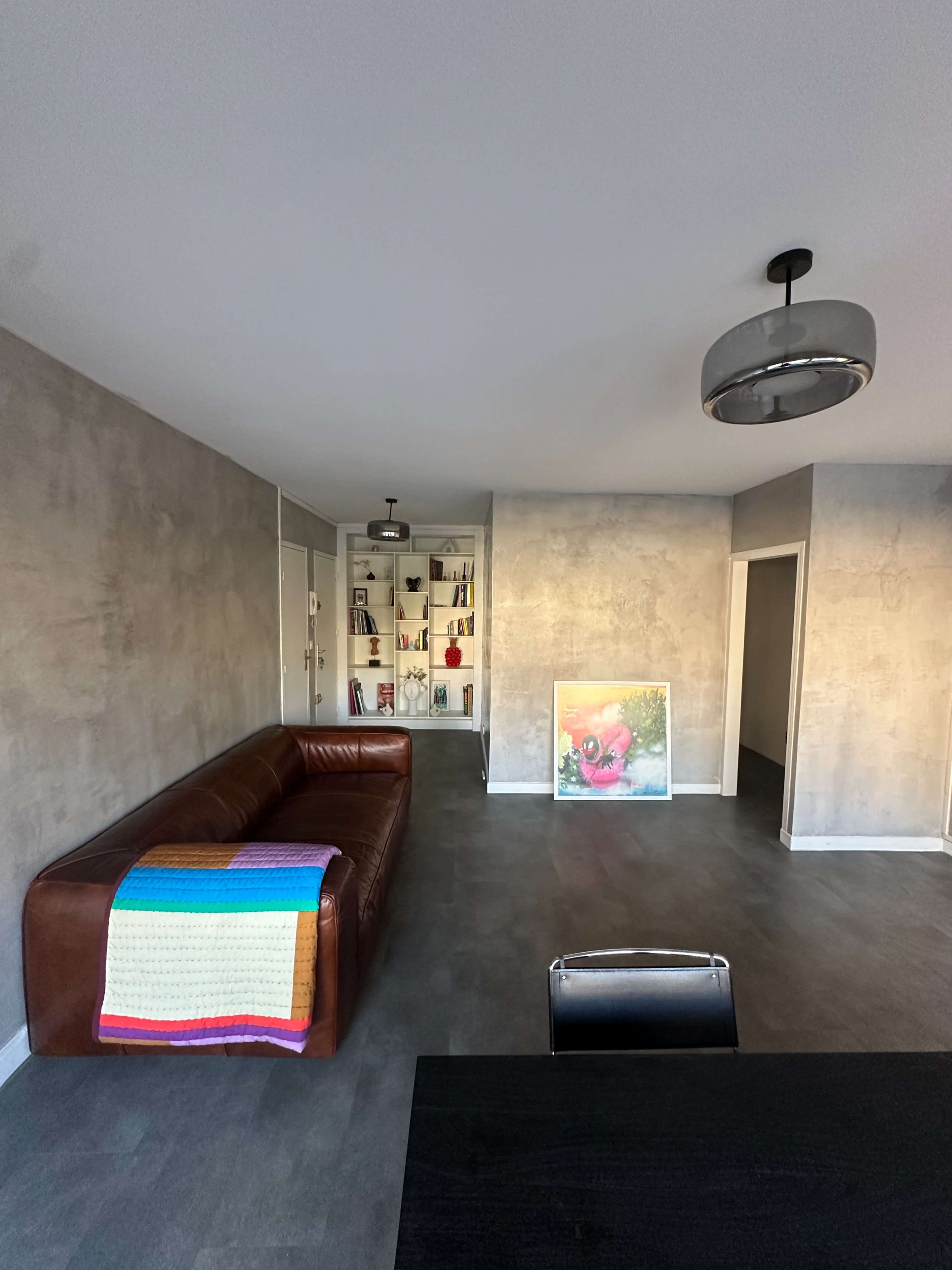 The image shows a modern living space with a brown leather couch, a colorful blanket draped over it, and a shelf filled with various items in the background.