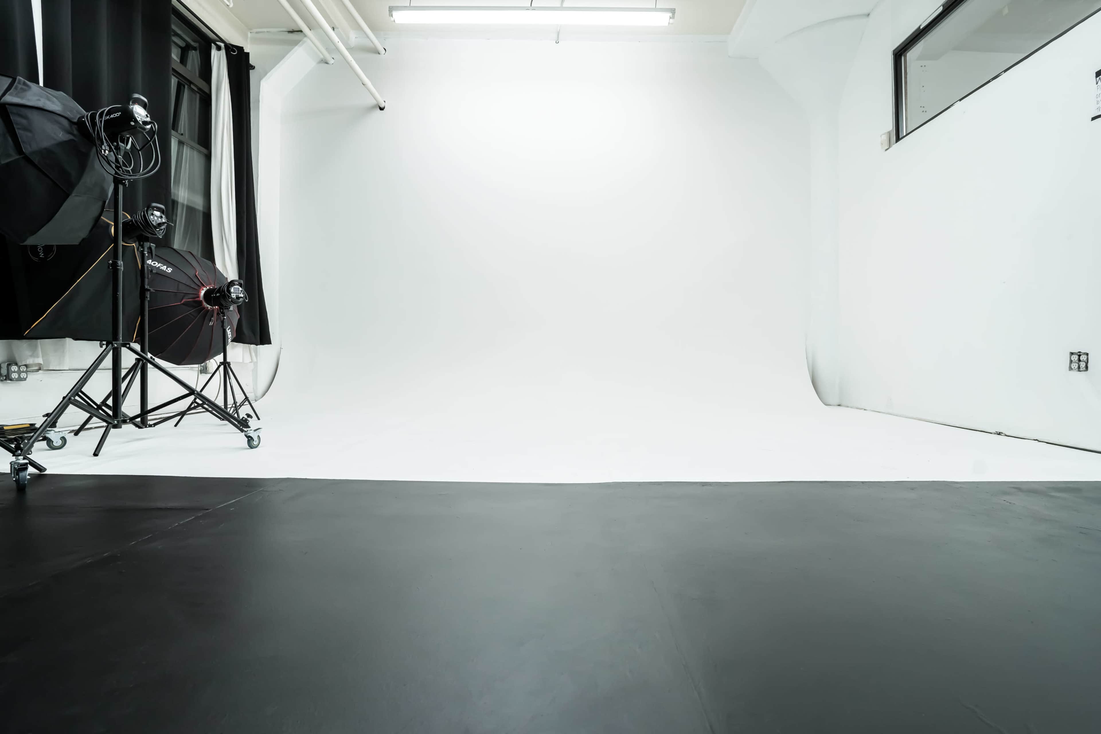 The image shows a photography studio with a white backdrop and a black floor, equipped with lighting and equipment on the left.