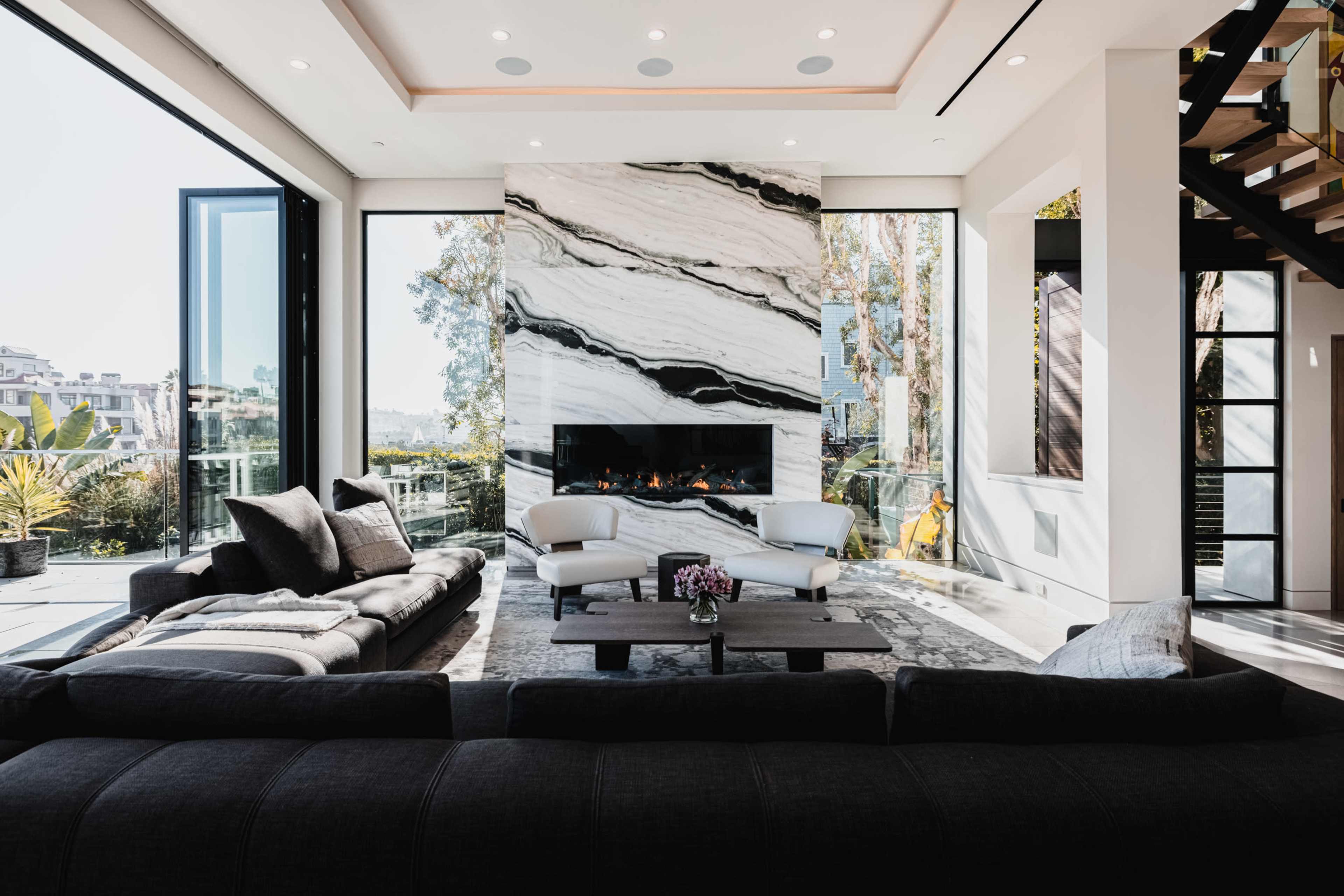 A modern living room features a large marble fireplace, floor-to-ceiling windows, and a sectional sofa arranged around a coffee table.
