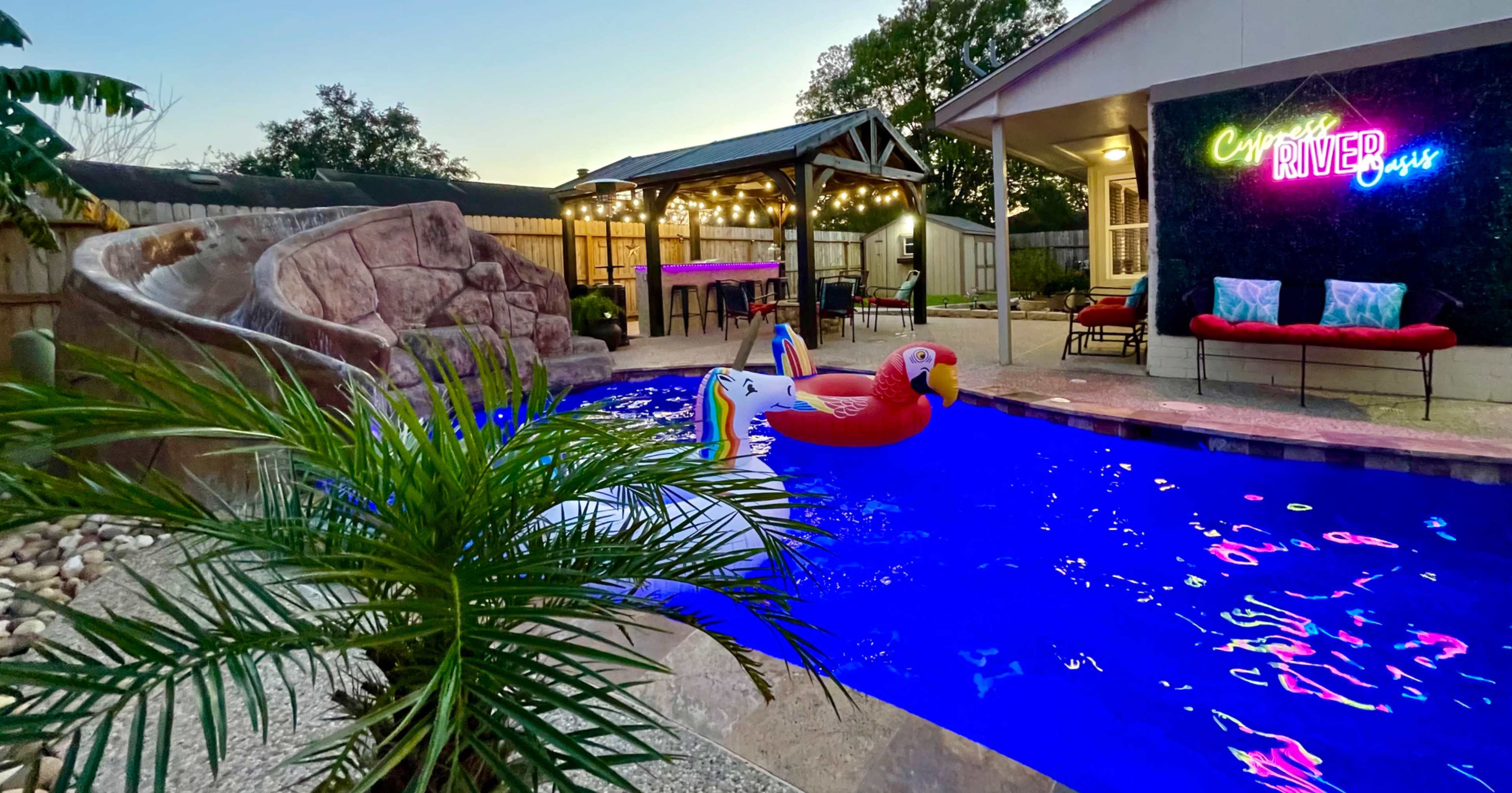 The scene shows a colorful swimming pool with a flamingo float, surrounded by tropical plants, a slide, and a covered outdoor seating area illuminated by string lights.
