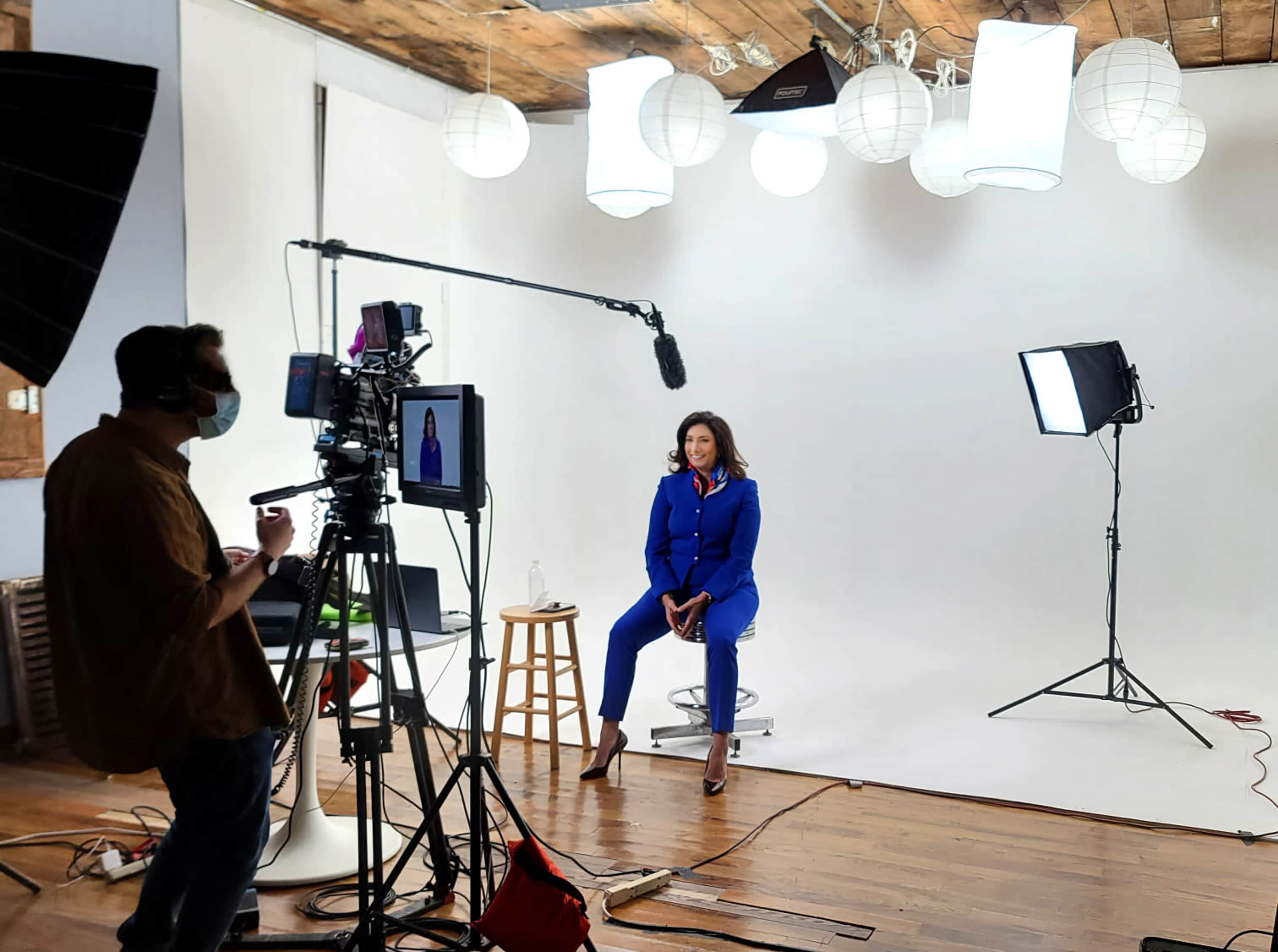 A woman in a blue suit sits on a stool in front of a camera setup in a brightly lit studio with softbox lights and paper lanterns overhead.
