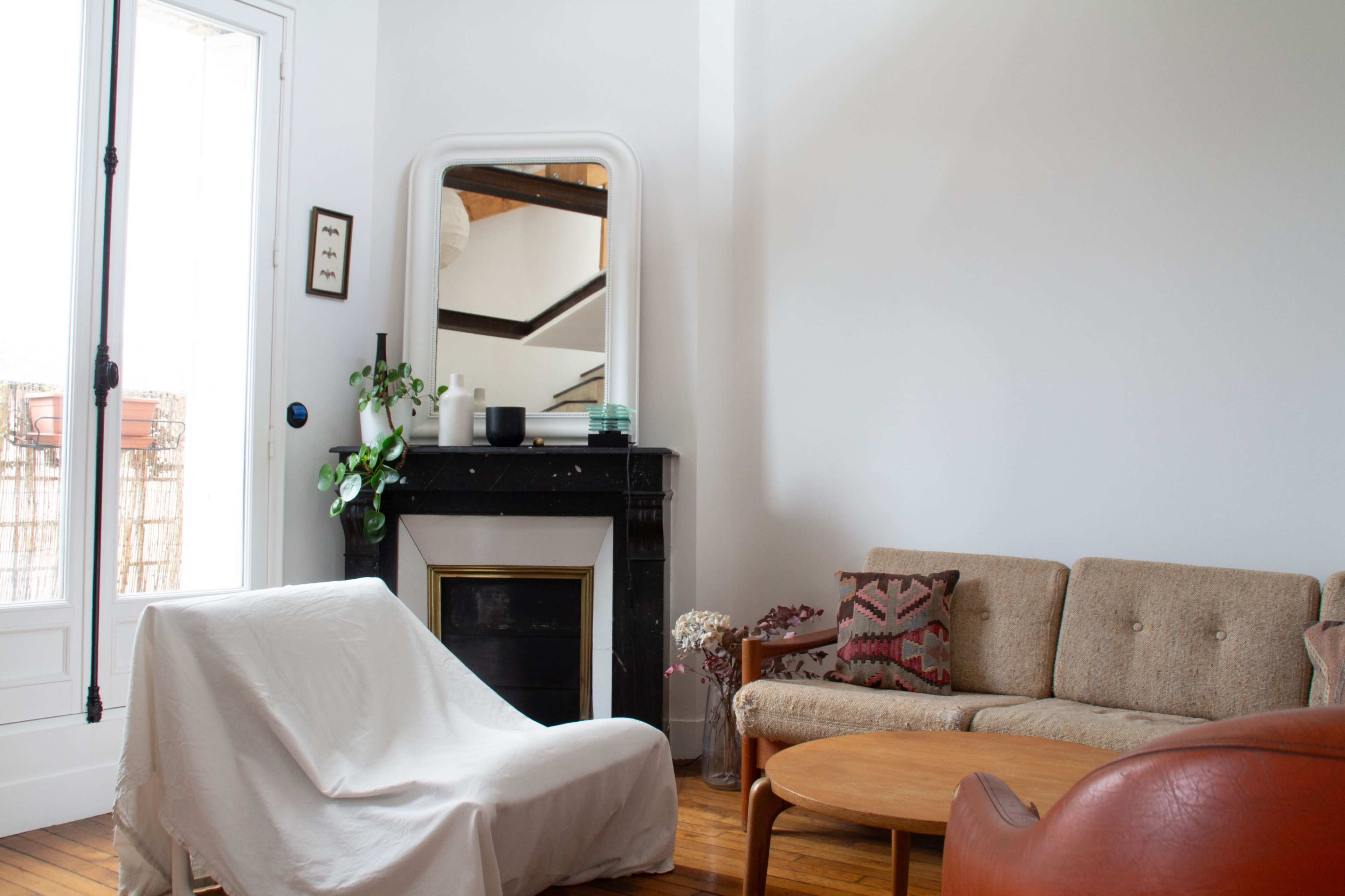 A living room with a sofa, a chair, a round table, and a fireplace, featuring a large mirror and a potted plant near a window.