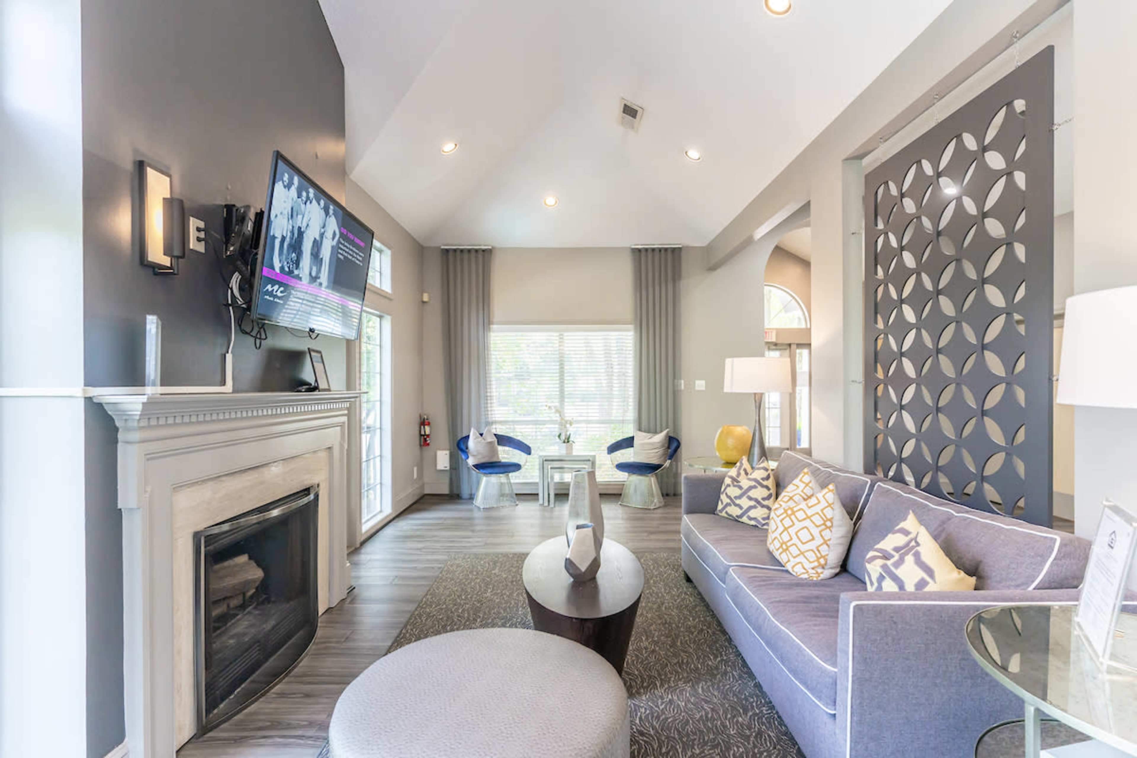 A modern living room with a gray sofa, a round coffee table, a fireplace, and a television mounted on the wall, featuring large windows that provide natural light.