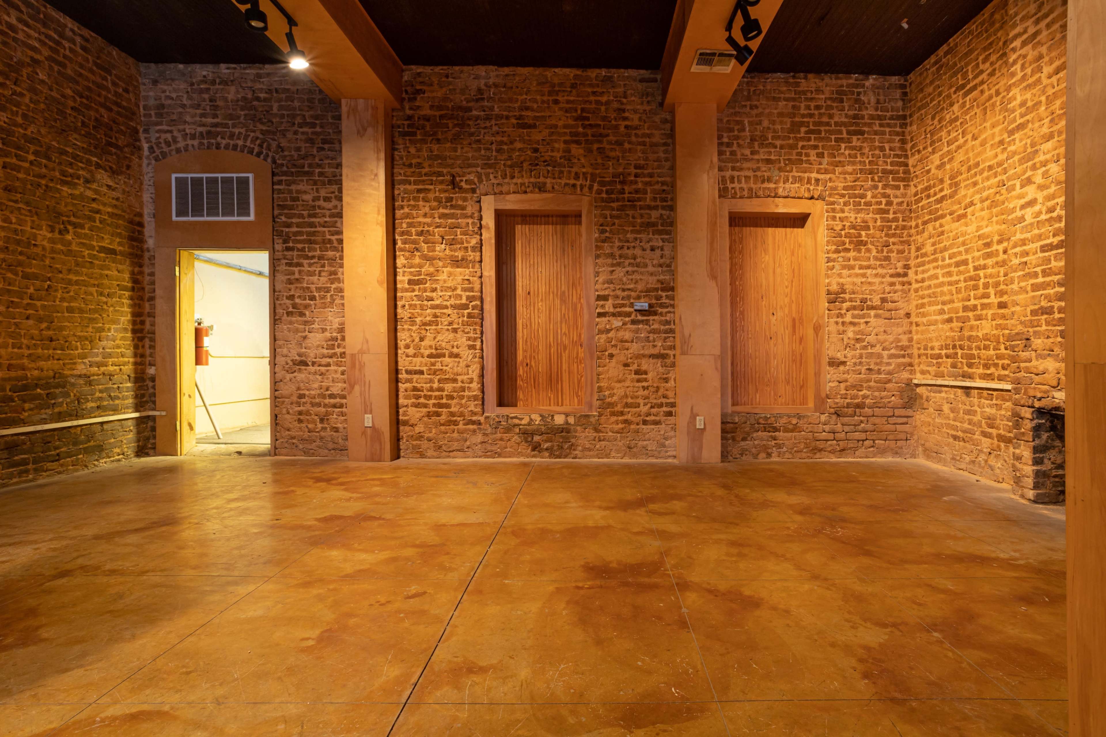 The image shows an empty room with exposed brick walls and a polished concrete floor, featuring two wooden panel wall recesses and a doorway leading to another space.