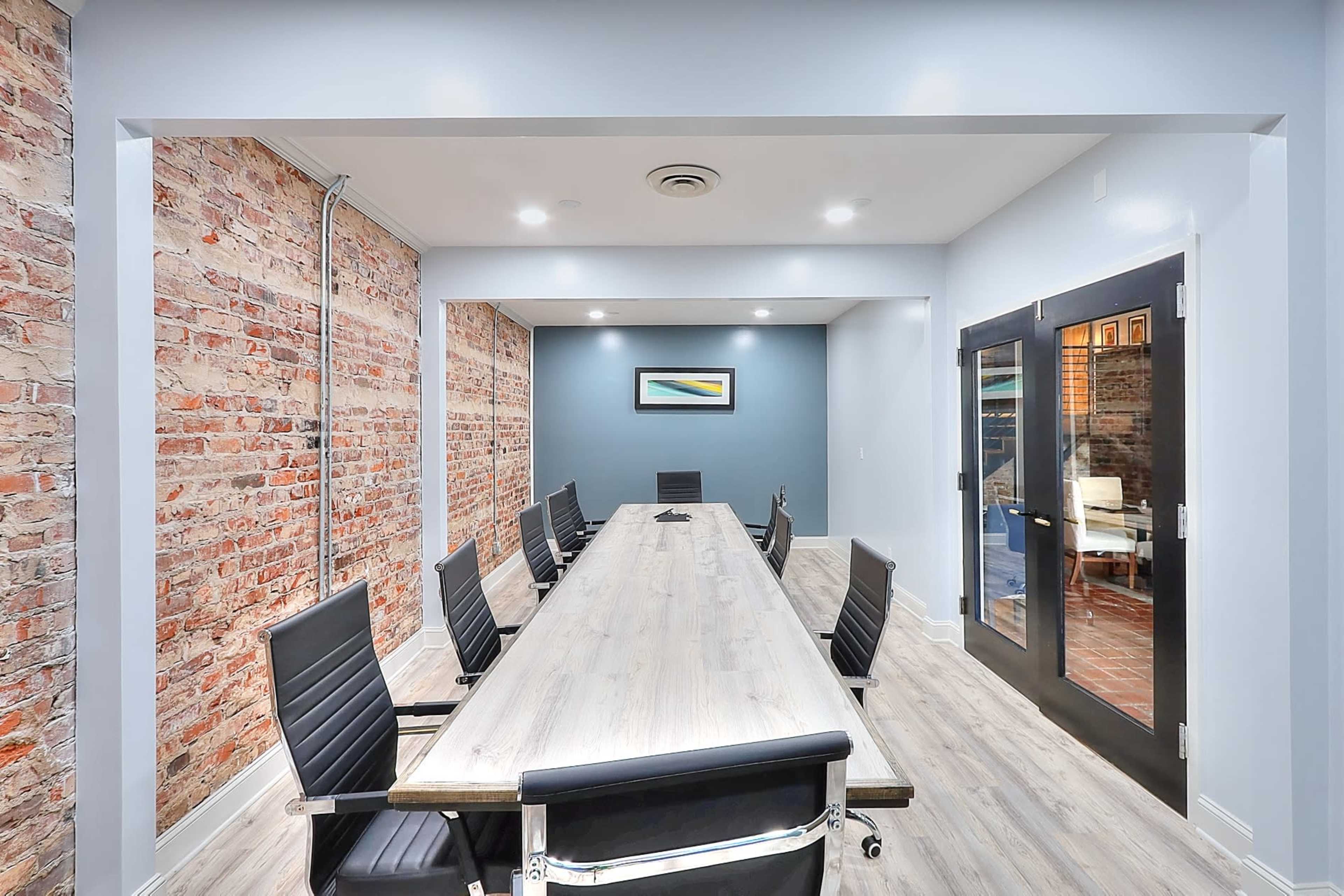 A conference room with a long wooden table surrounded by black chairs, featuring exposed brick walls and modern lighting.